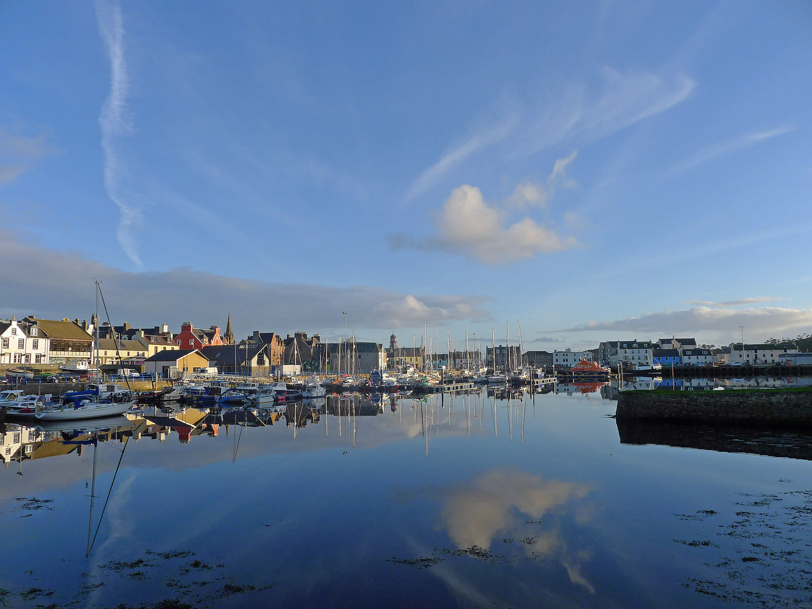 A calm harbour scene in Stornoway with several small boats moored, reflecting the blue sky and clouds on the still water surface, with buildings lining the shore in the background.