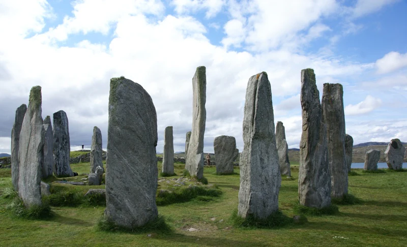 A group of tall, weathered standing stones arranged in a circular formation on a grassy field under a partly cloudy sky.