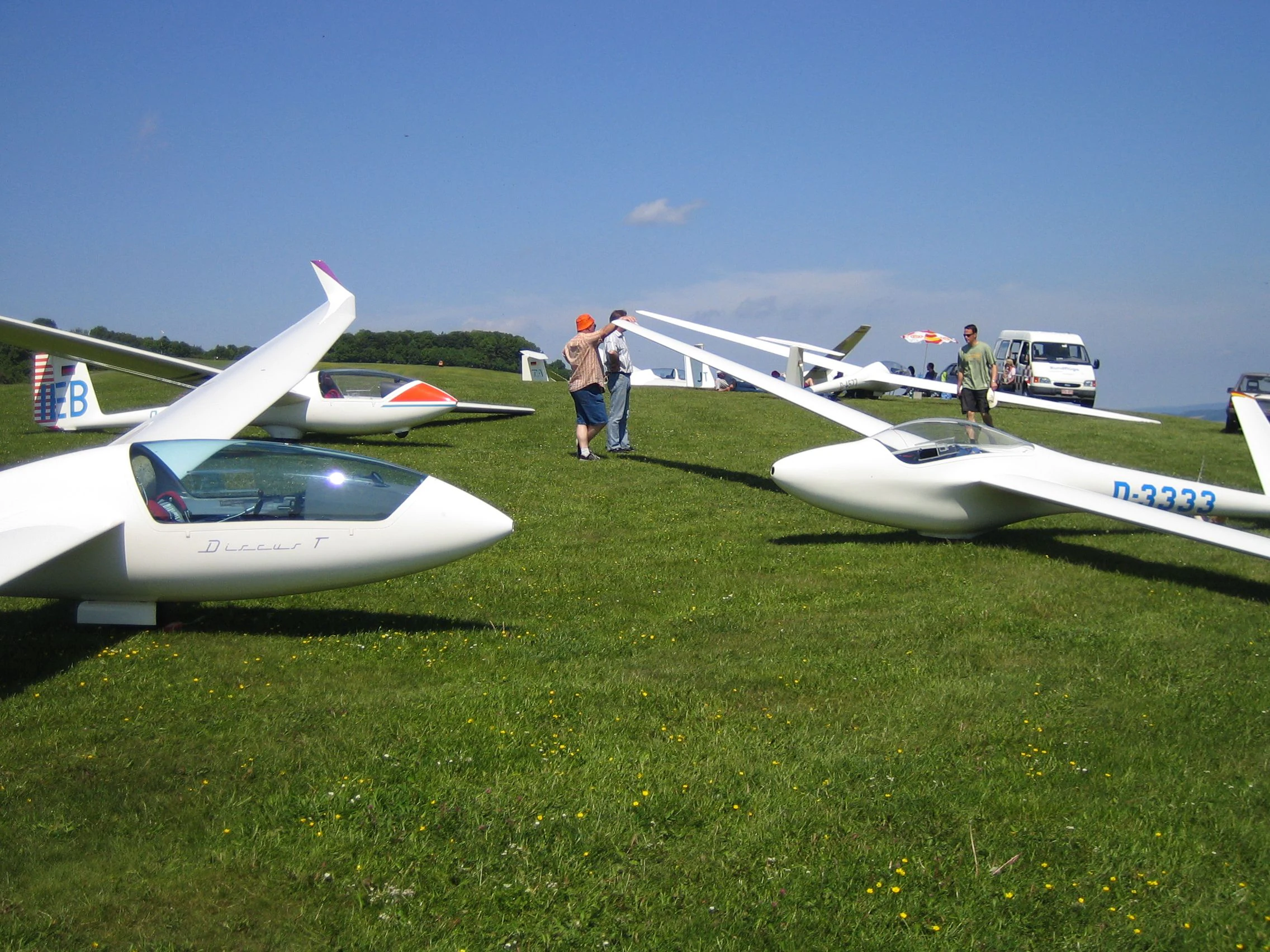 Several white gliders parked on a wide green meadow under a clear blue sky, with people standing nearby and a van in the background.