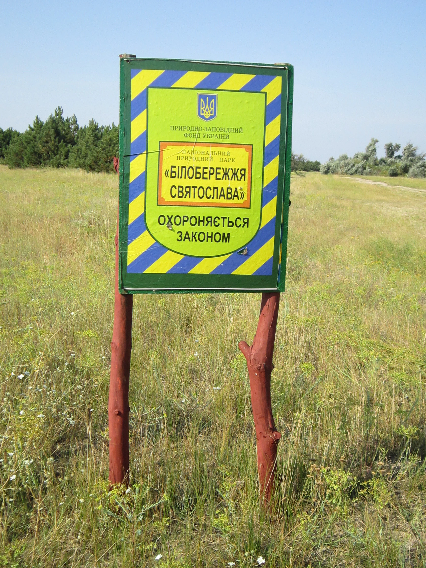 Green park sign with yellow and blue stripes displaying Ukrainian text mounted on two wooden posts in a grassy field