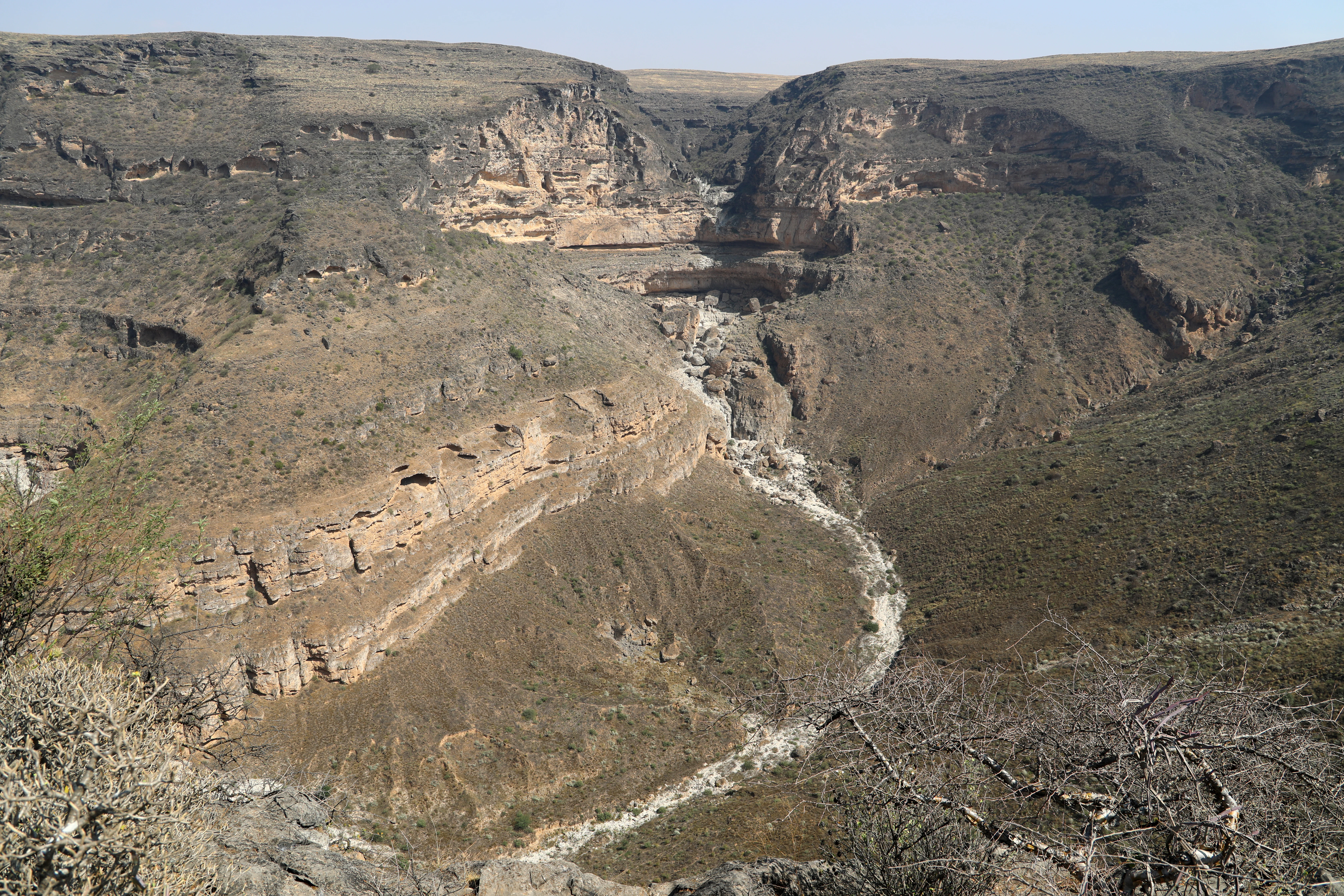 A wide, high-angle view looking down into a deep, rocky sinkhole crater with layered canyon walls and sparse desert vegetation.