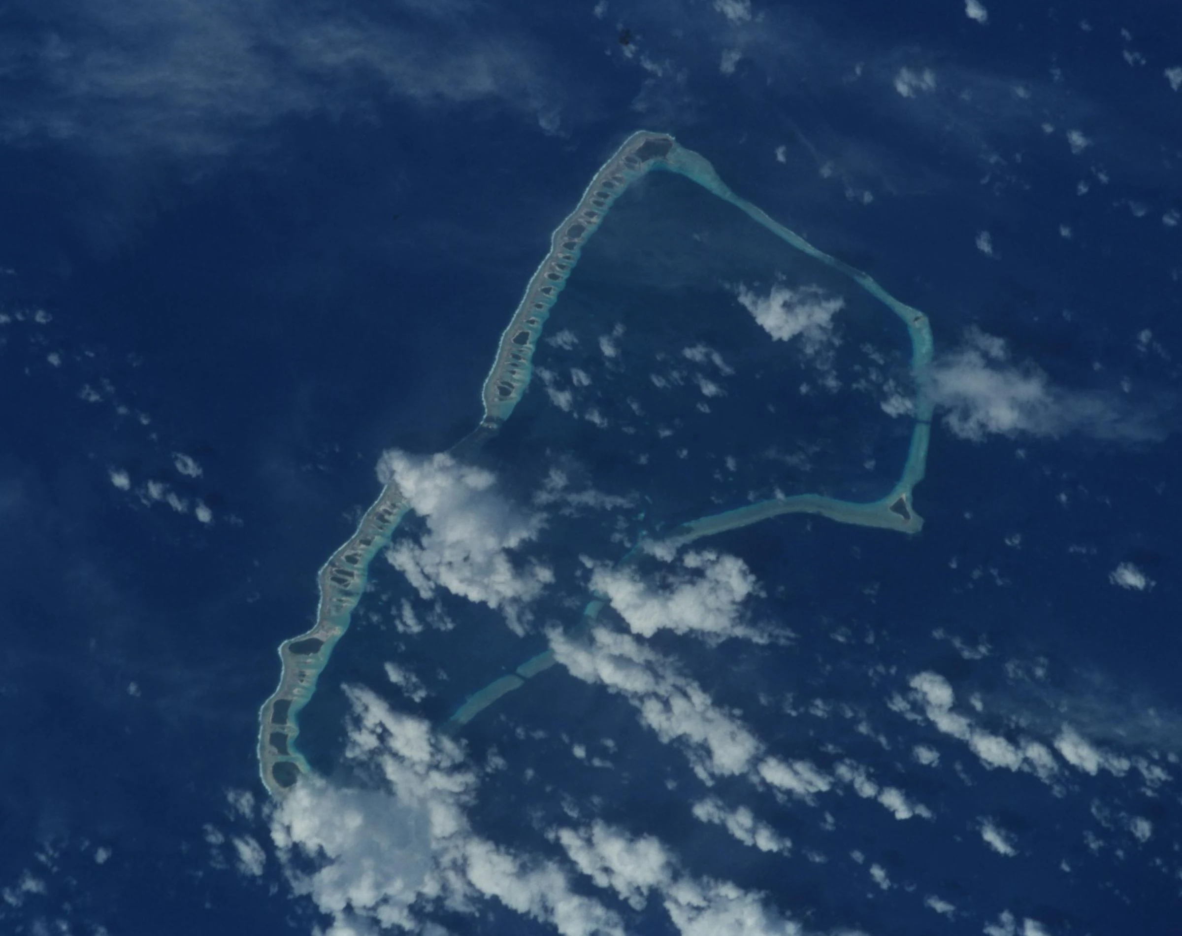 An aerial satellite photograph of Jaluit Atoll, showing a ring of coral islets surrounded by the deep blue Pacific Ocean with scattered white clouds.