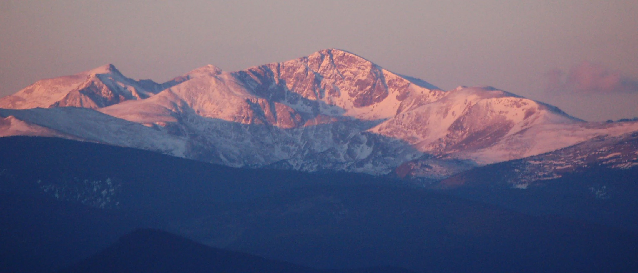 A panoramic, wide-angle view of James Peak covered in snow, glowing with golden-pink morning light, rising above dark, shadowed mountain foothills.