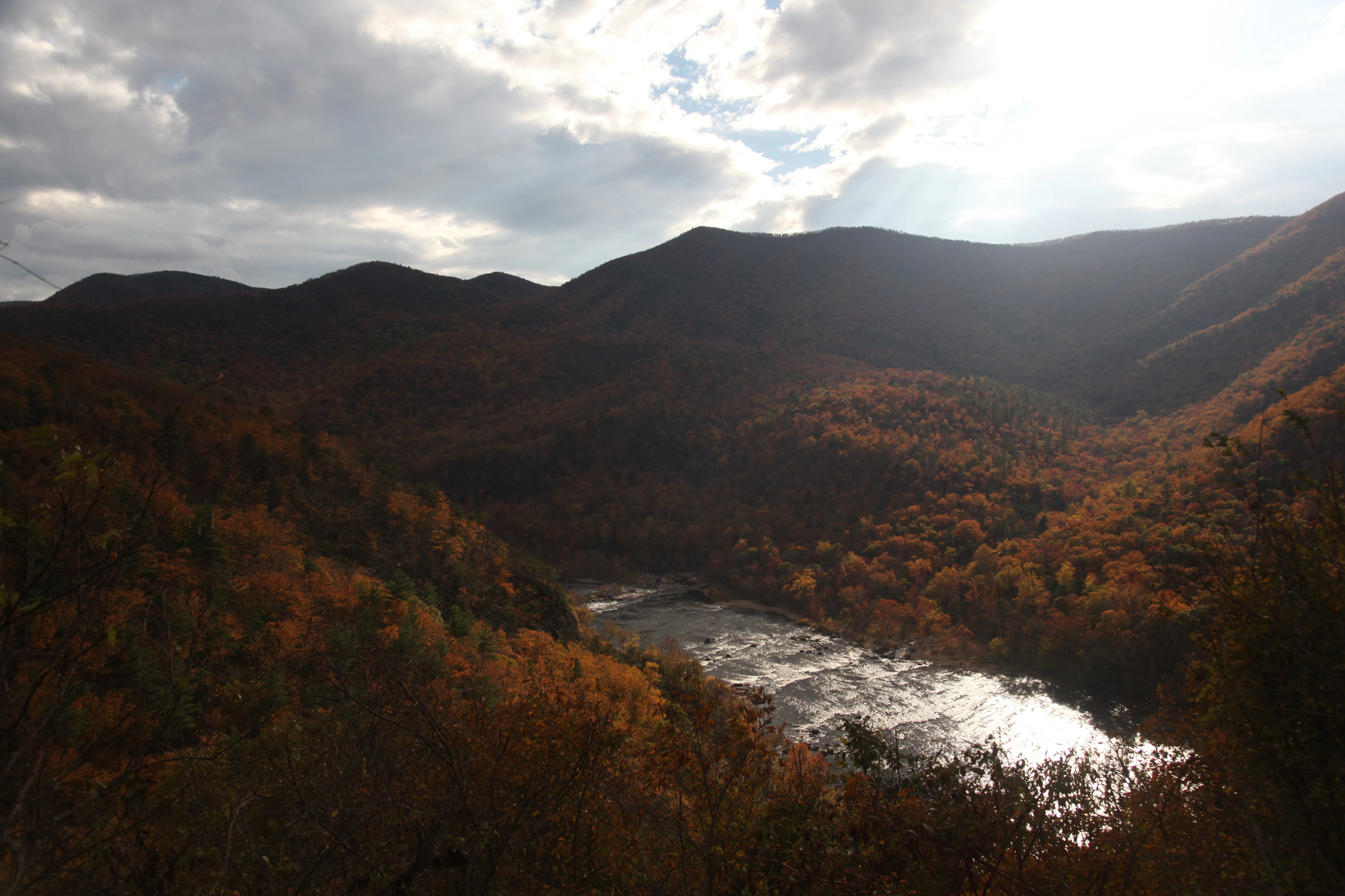 A mountain valley with a river reflecting sunlight, surrounded by hills covered in autumn-colored trees under a cloudy sky with bright sun glare.