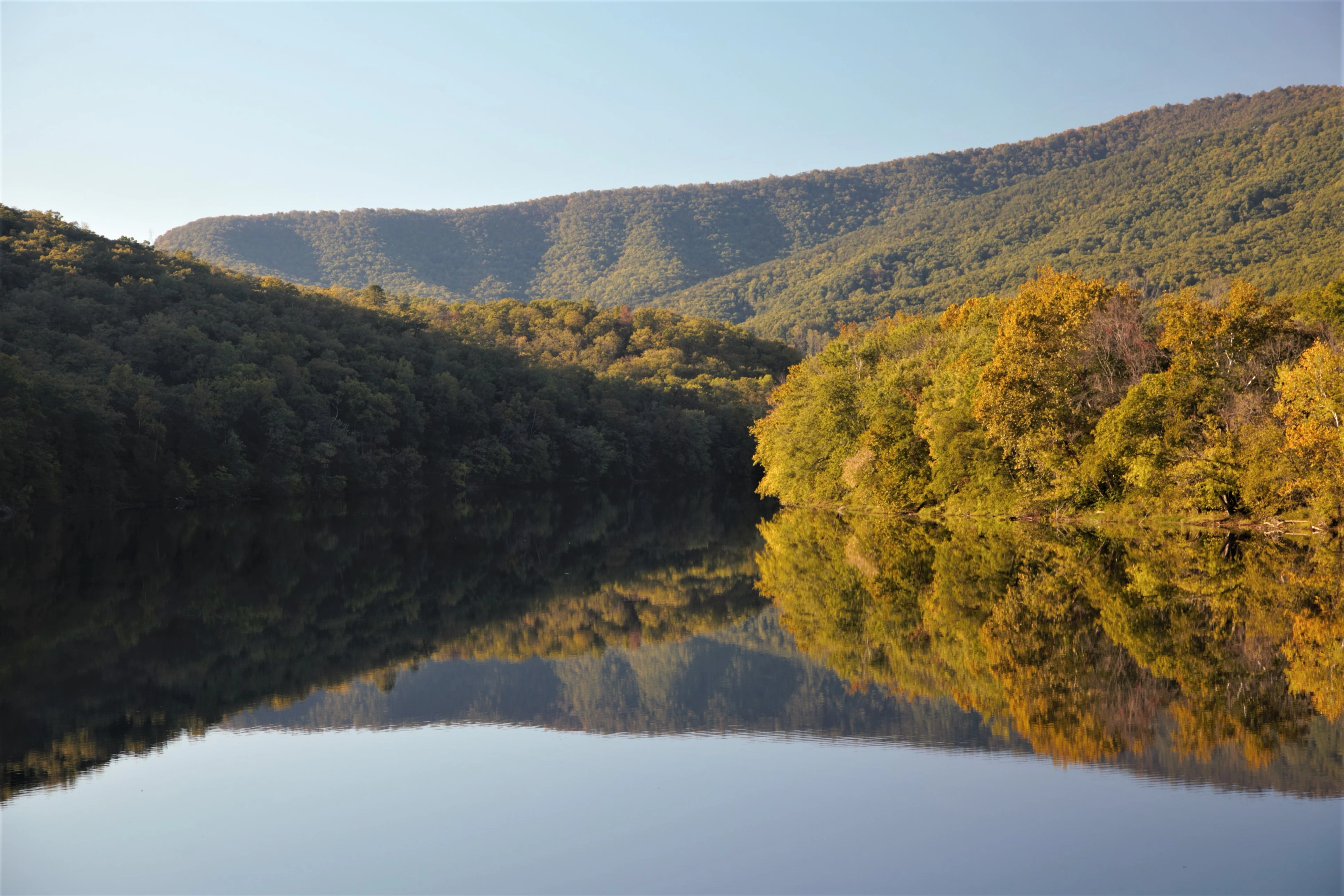 A calm river reflecting forested hills with autumn foliage under a clear blue sky.