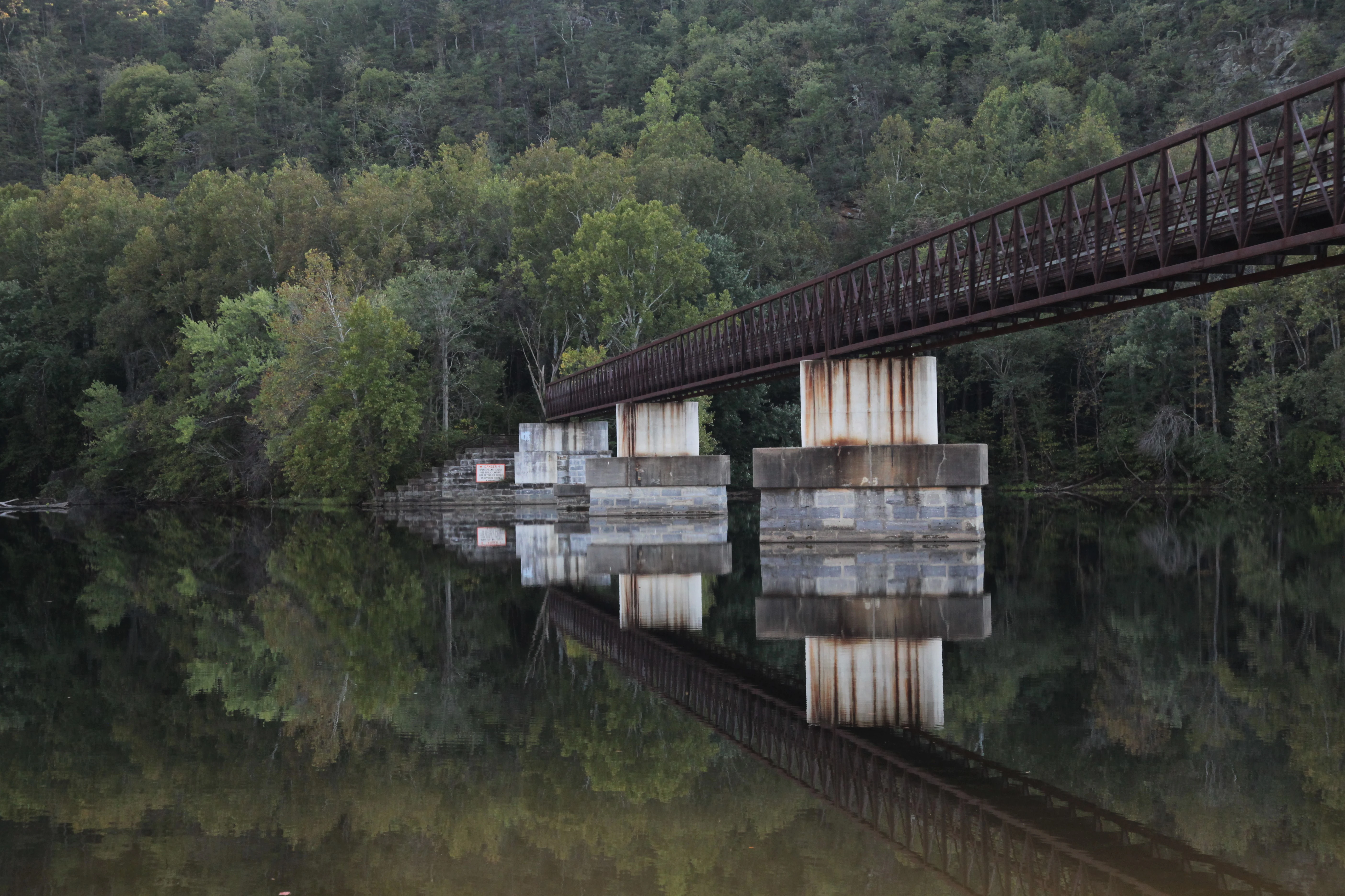 A metal footbridge with concrete piers spans a calm river, with the bridge structure and surrounding forest reflected in the still water below