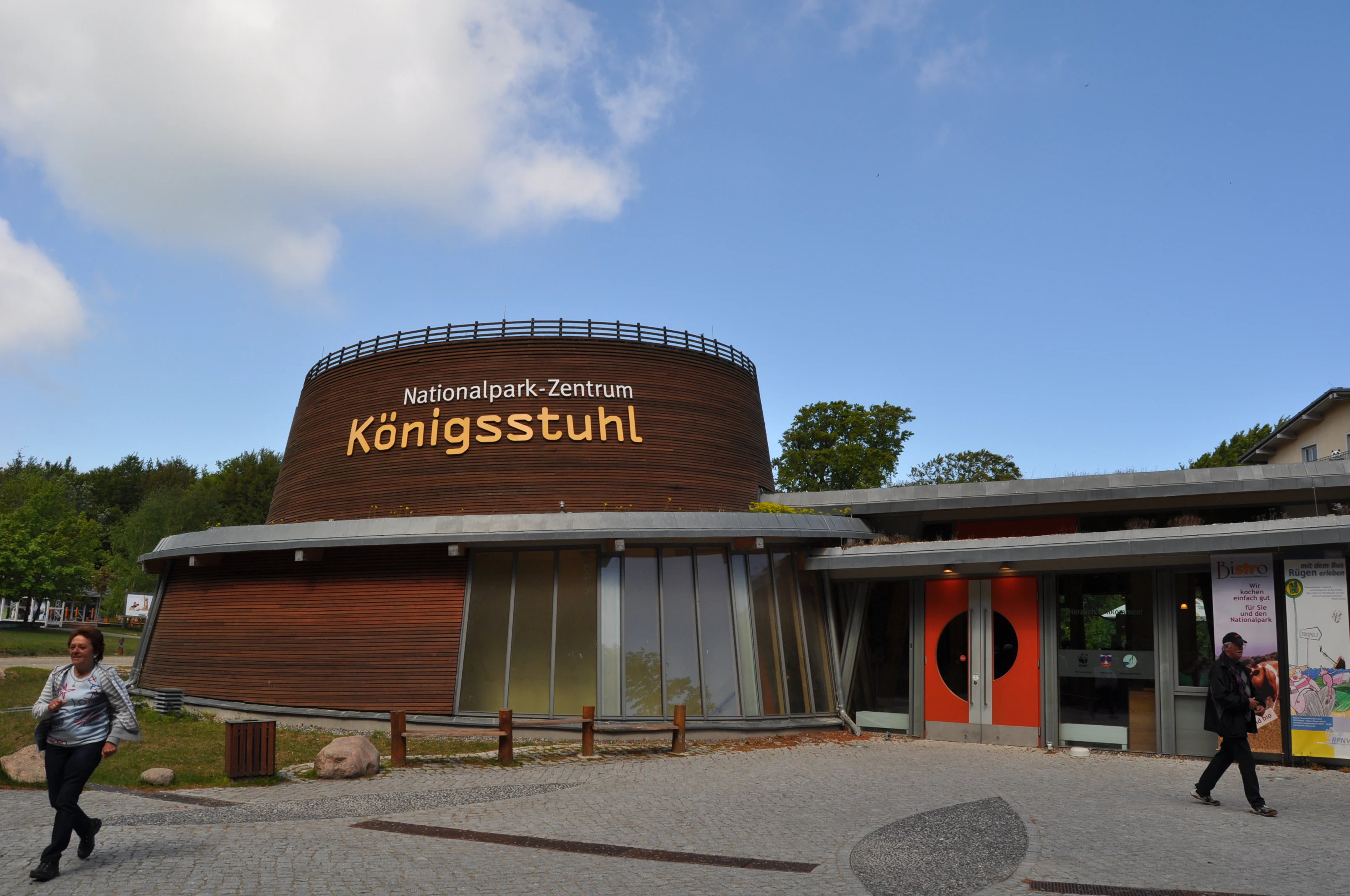 A modern building labeled 'Nationalpark-Zentrum Königsstuhl' with a curved brown roof, glass facade, and orange doors, surrounded by trees and a paved area with people walking