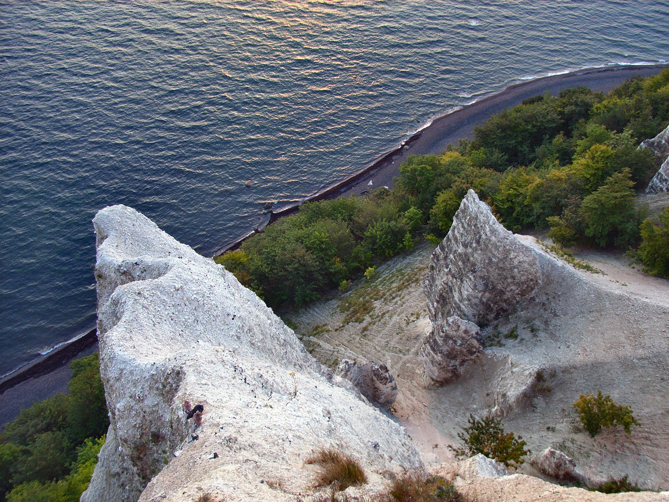 White chalk cliffs overlooking the sea with forested coastline below and a small figure of a person on the cliff face