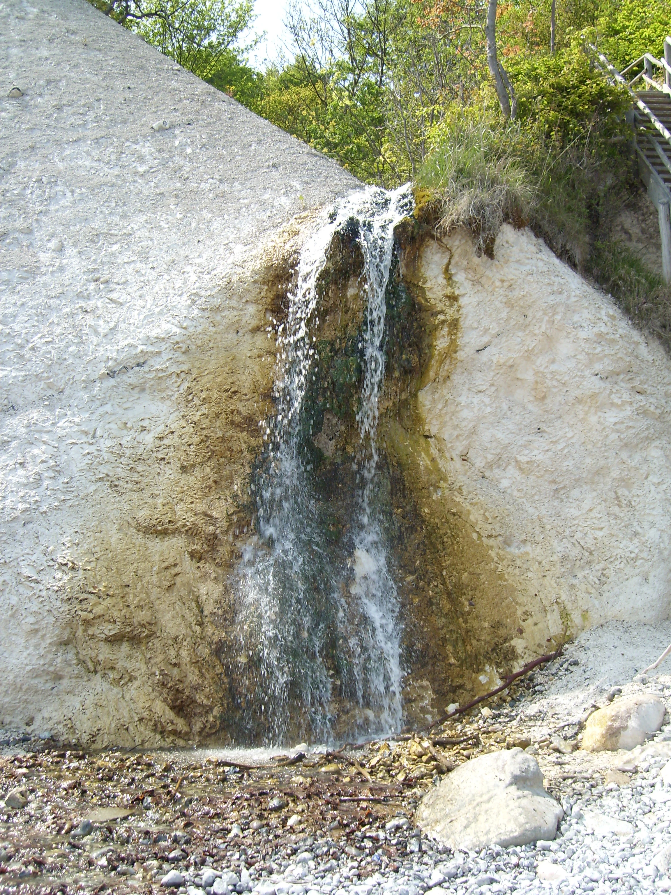 Waterfall flowing down a light-colored chalk cliff with rocks and pebbles on the ground below, surrounded by greenery at the cliff top
