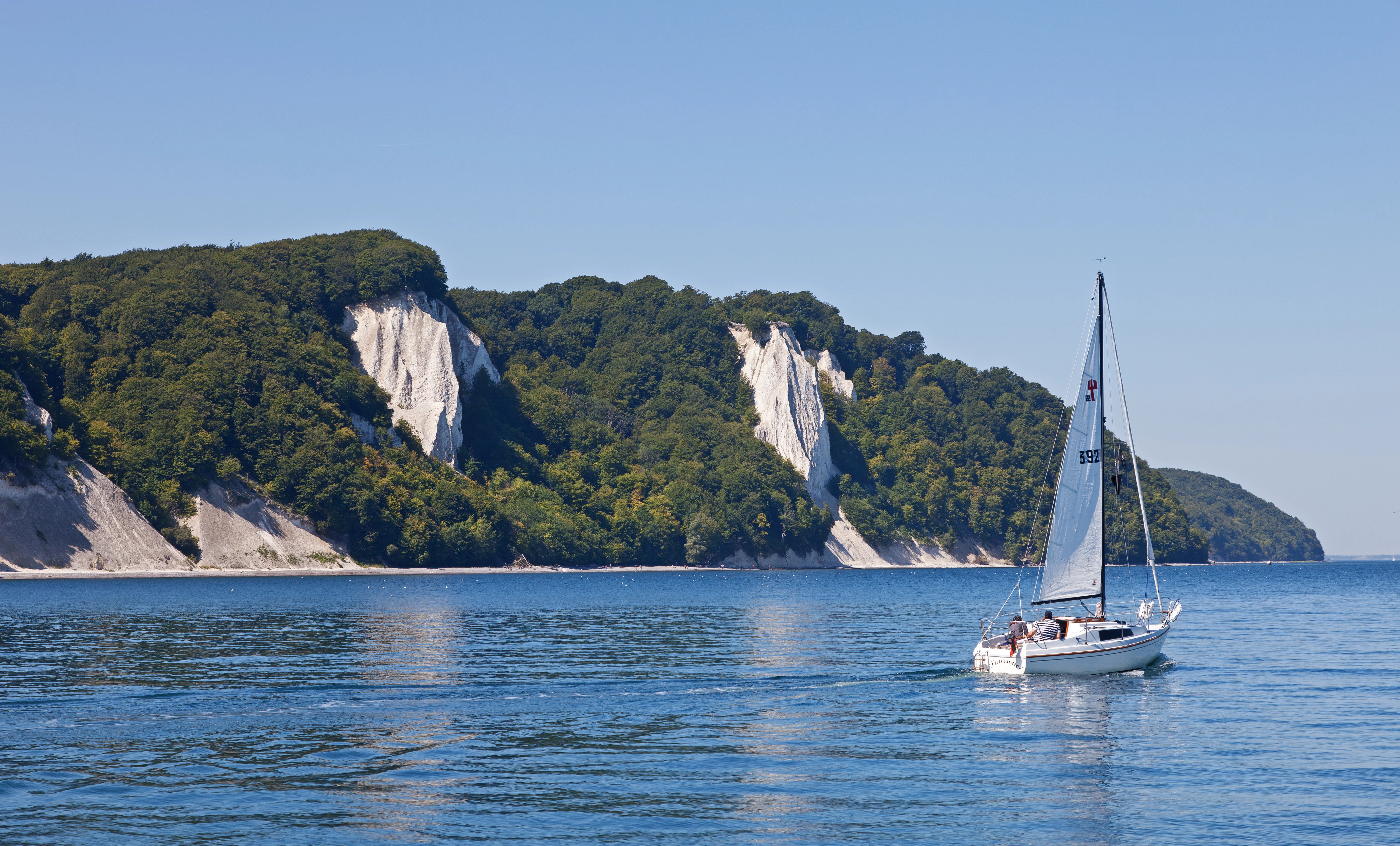 A sailboat on calm blue water with white chalk cliffs covered in green trees under a clear blue sky