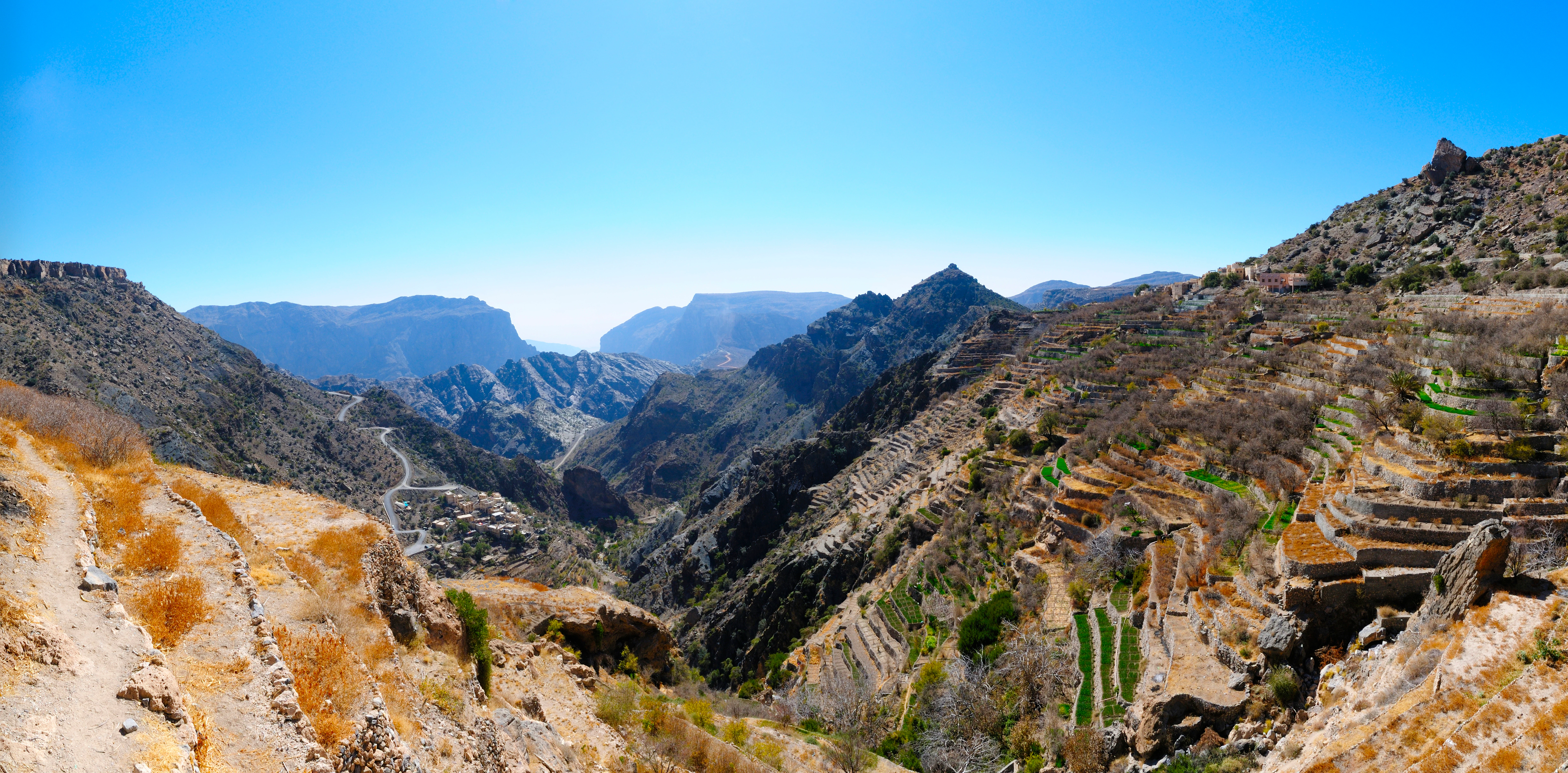 A wide landscape view of terraced mountains in the Hajar range, featuring stone-walled fields built into steep, rocky terrain with a deep valley between peaks under a clear blue sky.