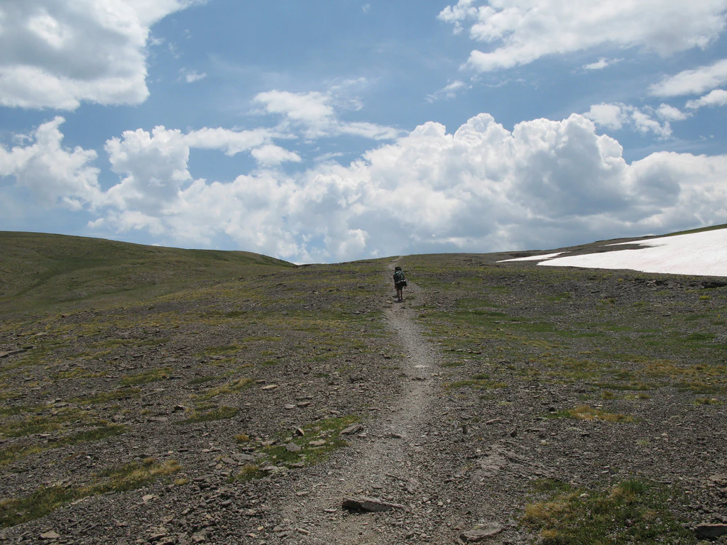 A lone hiker walks along a narrow, rocky dirt trail winding through a barren, high-alpine landscape under a bright, cloudy sky with a patch of snow visible on the right.