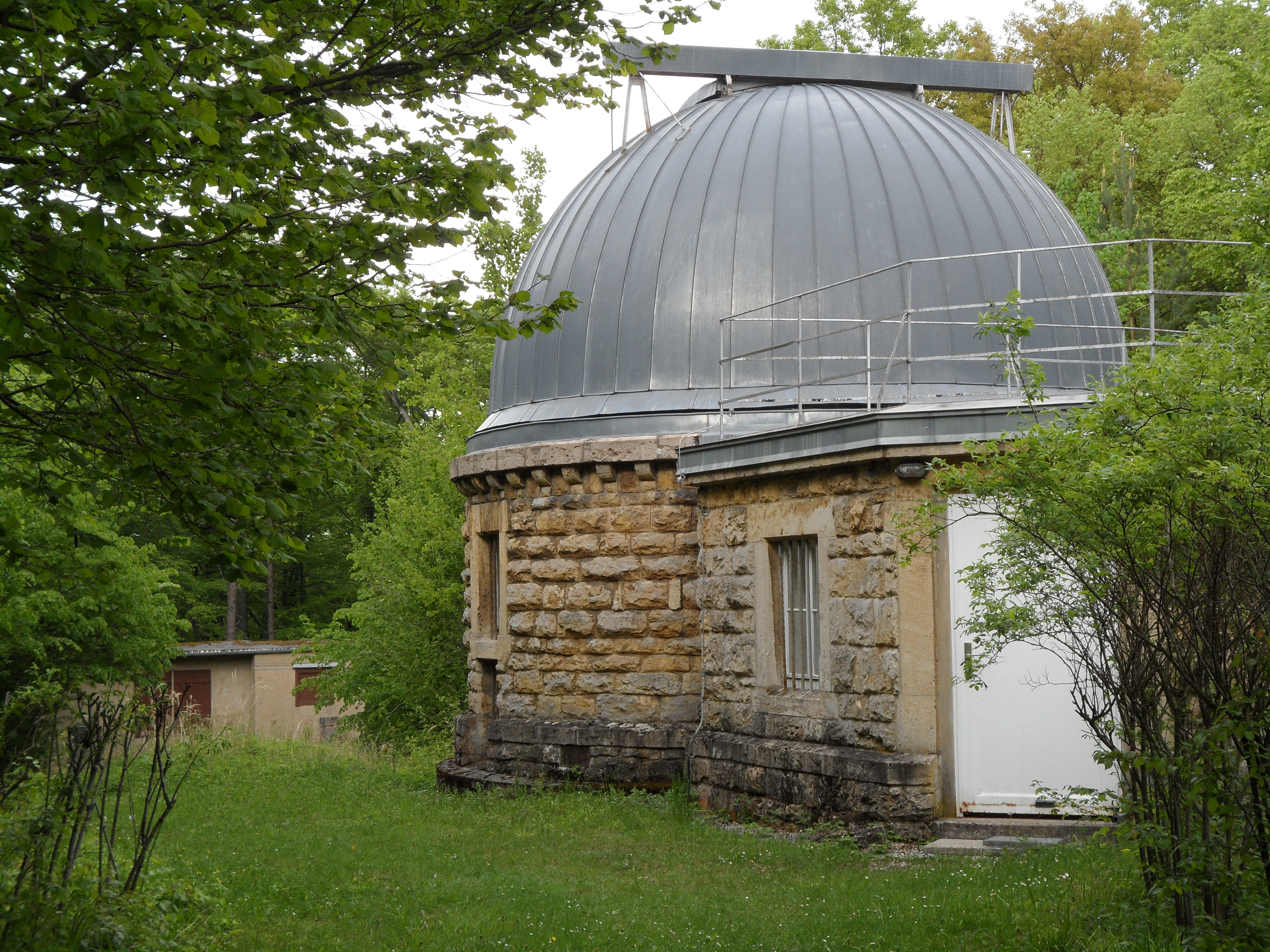A round, stone-built observatory with a metallic dome roof, surrounded by green trees and foliage in the Jenaer Forst.