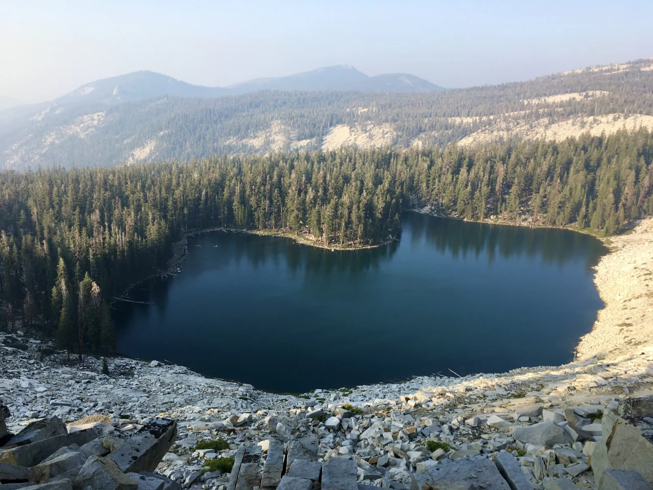 An elevated, wide view of a calm, dark-blue mountain lake surrounded by dense evergreen forest and steep, rocky, light-colored slopes.