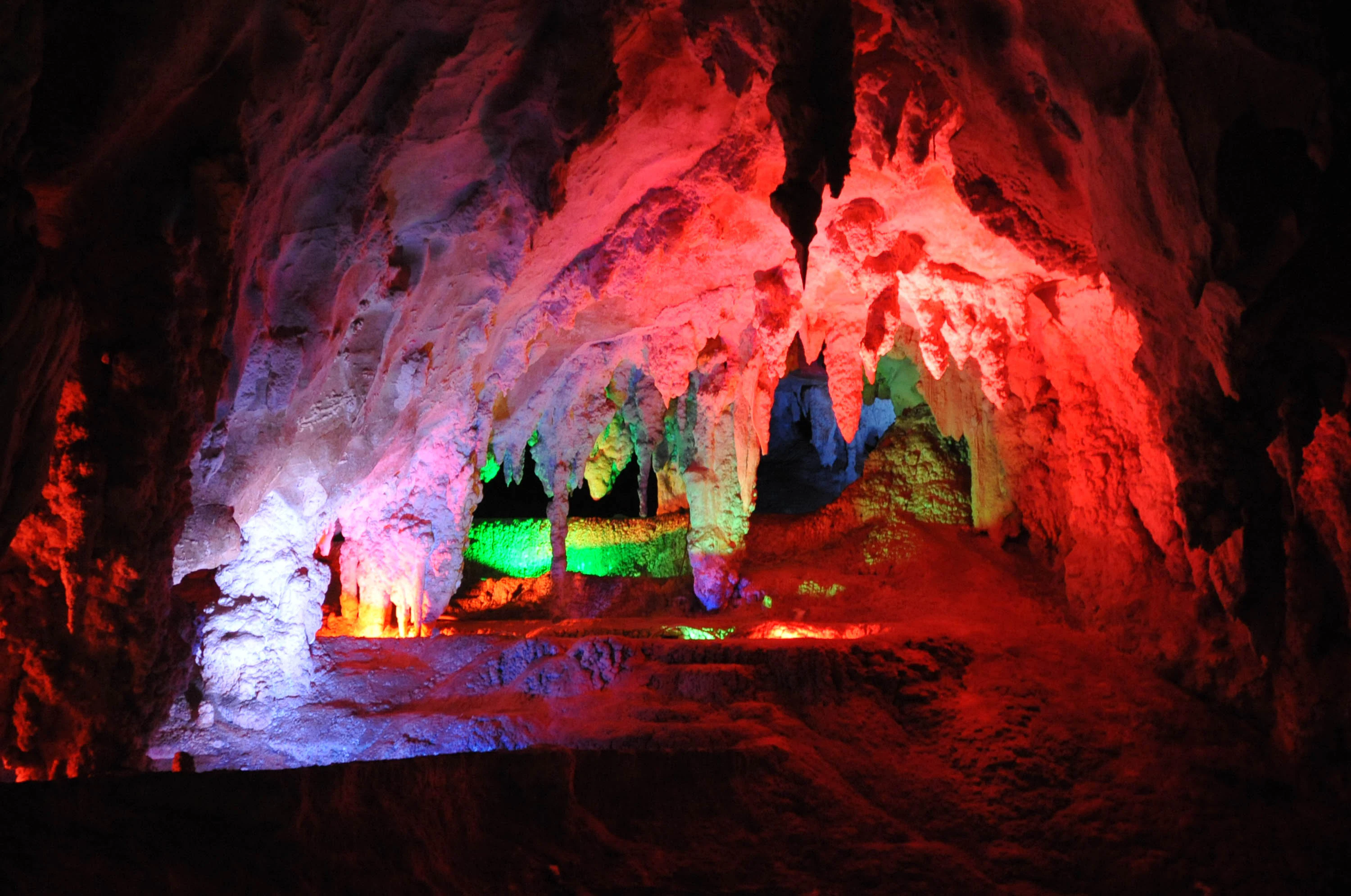 A cavern interior with limestone stalactites and stalagmites illuminated by vibrant red, green, and blue artificial lights.