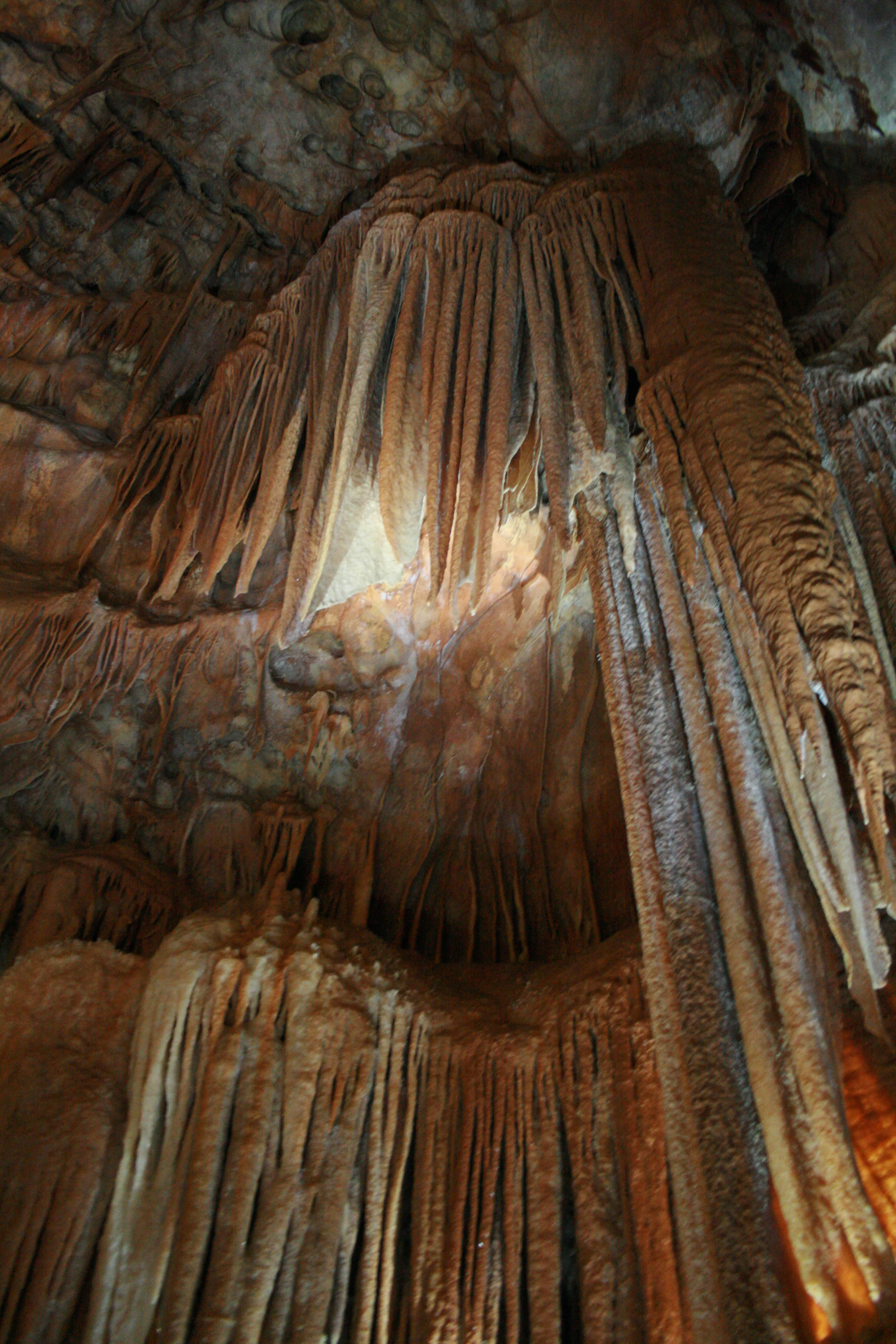 A collection of textured, brownish limestone stalactites and flowstone formations hanging from a cave ceiling.