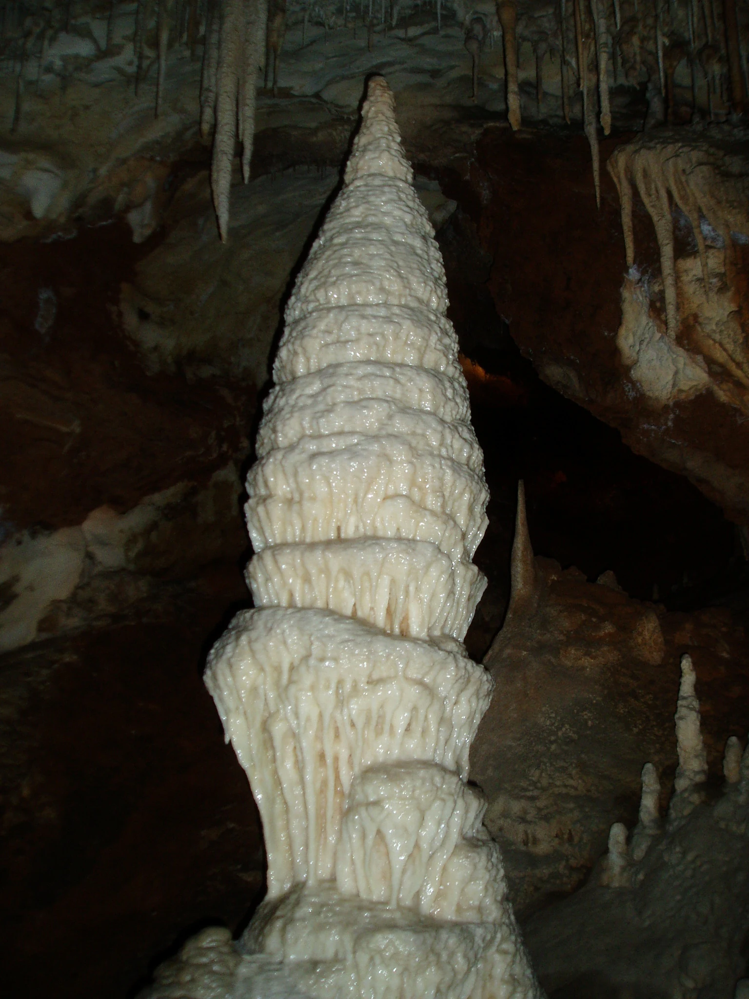 A large, white, cone-shaped stalagmite rising from the floor of a dark limestone cave, with smaller stalactites hanging from the ceiling above.