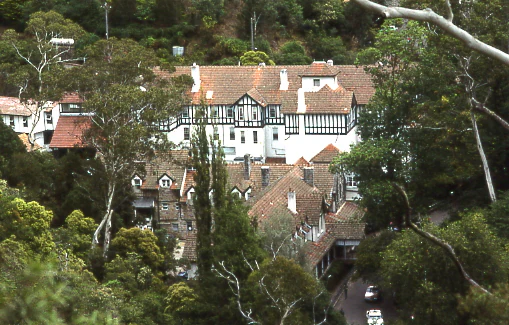 A wide, slightly elevated view of the historic, multi-story Caves House building, featuring white walls with black timber framing and brown tiled roofs, surrounded by a dense forest canopy.