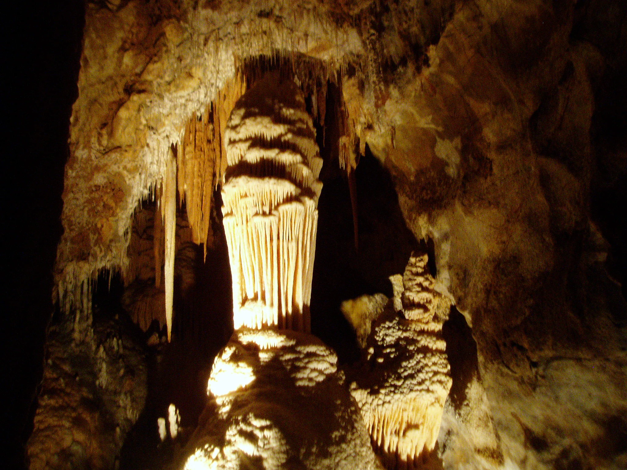 A large, illuminated central stalagmite column stands within a dark limestone cave, surrounded by smaller stalactites and straw formations hanging from the cavern ceiling.