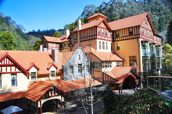 A large, multi-story historic hotel building with half-timbered walls, orange tile roofs, and stone accents, set against a backdrop of forested hills.