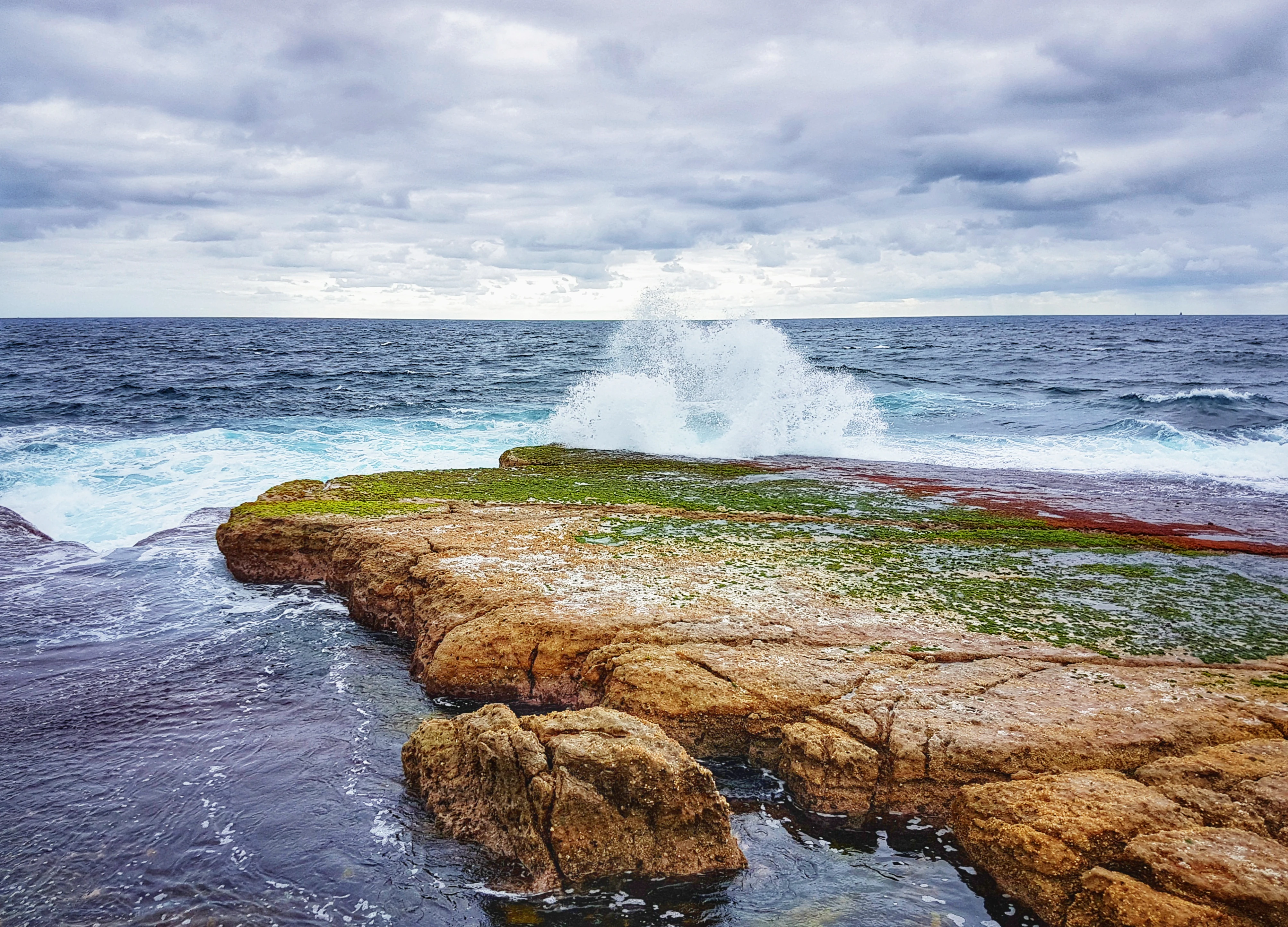 Ocean waves break against a flat, moss-covered rock formation extending into the sea under a cloudy sky.