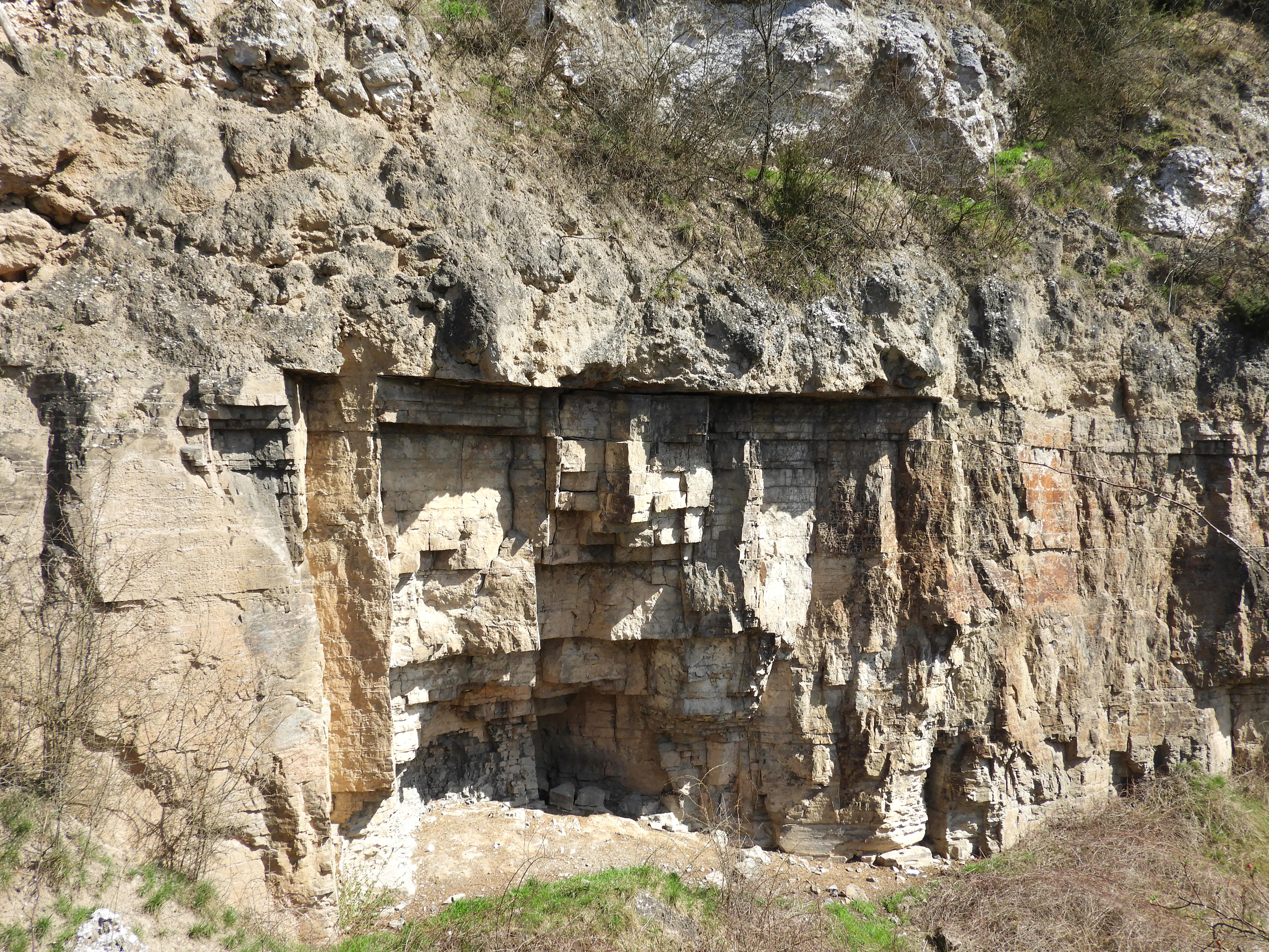 A steep rock face displaying horizontal geological strata in shades of grey, white, and brown, with sparse green vegetation at the top and bottom.