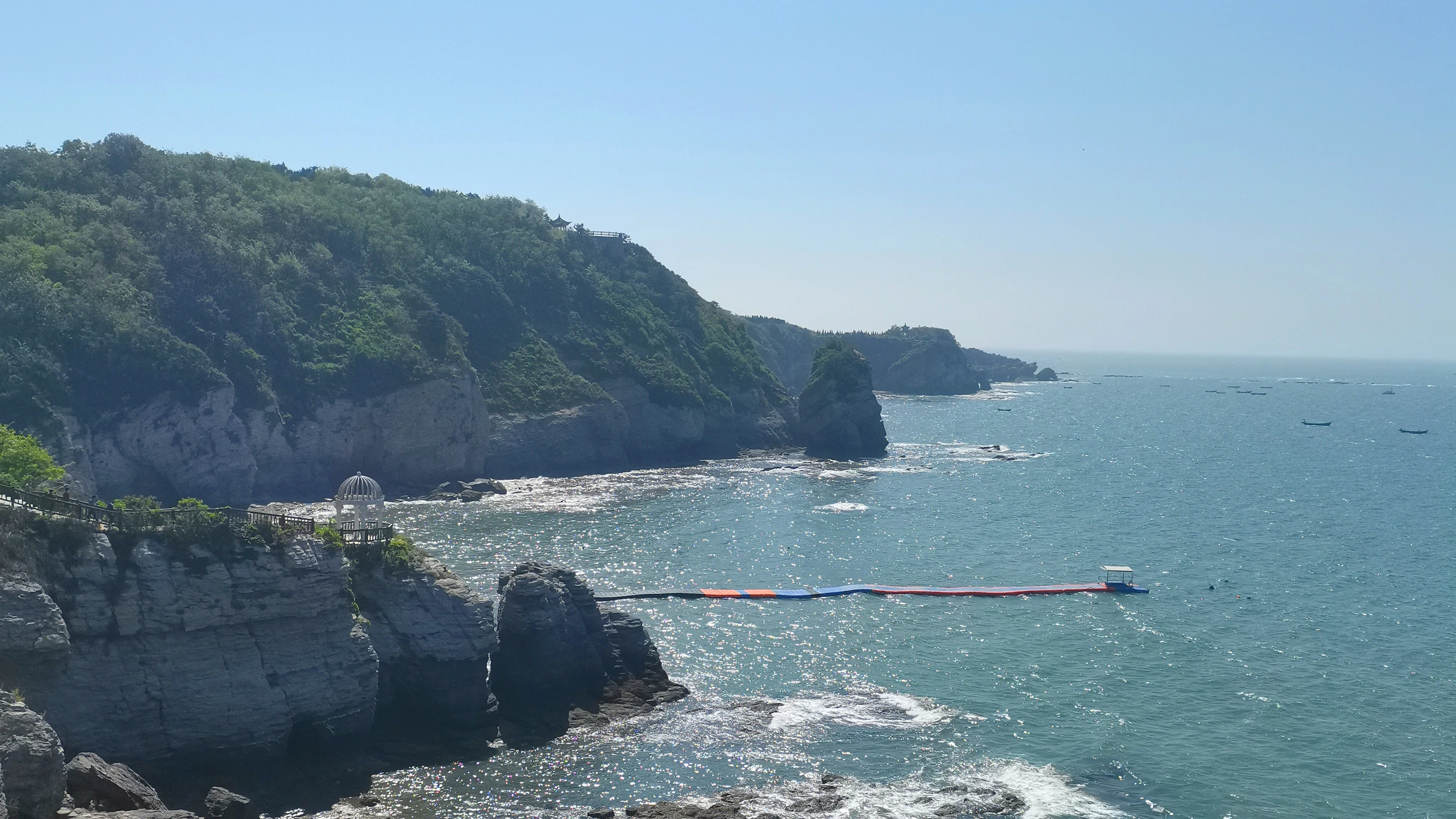 A wide coastal view of Jinshitan National Resort, featuring steep rocky cliffs topped with green vegetation overlooking the ocean, with a long floating walkway extending into the water.
