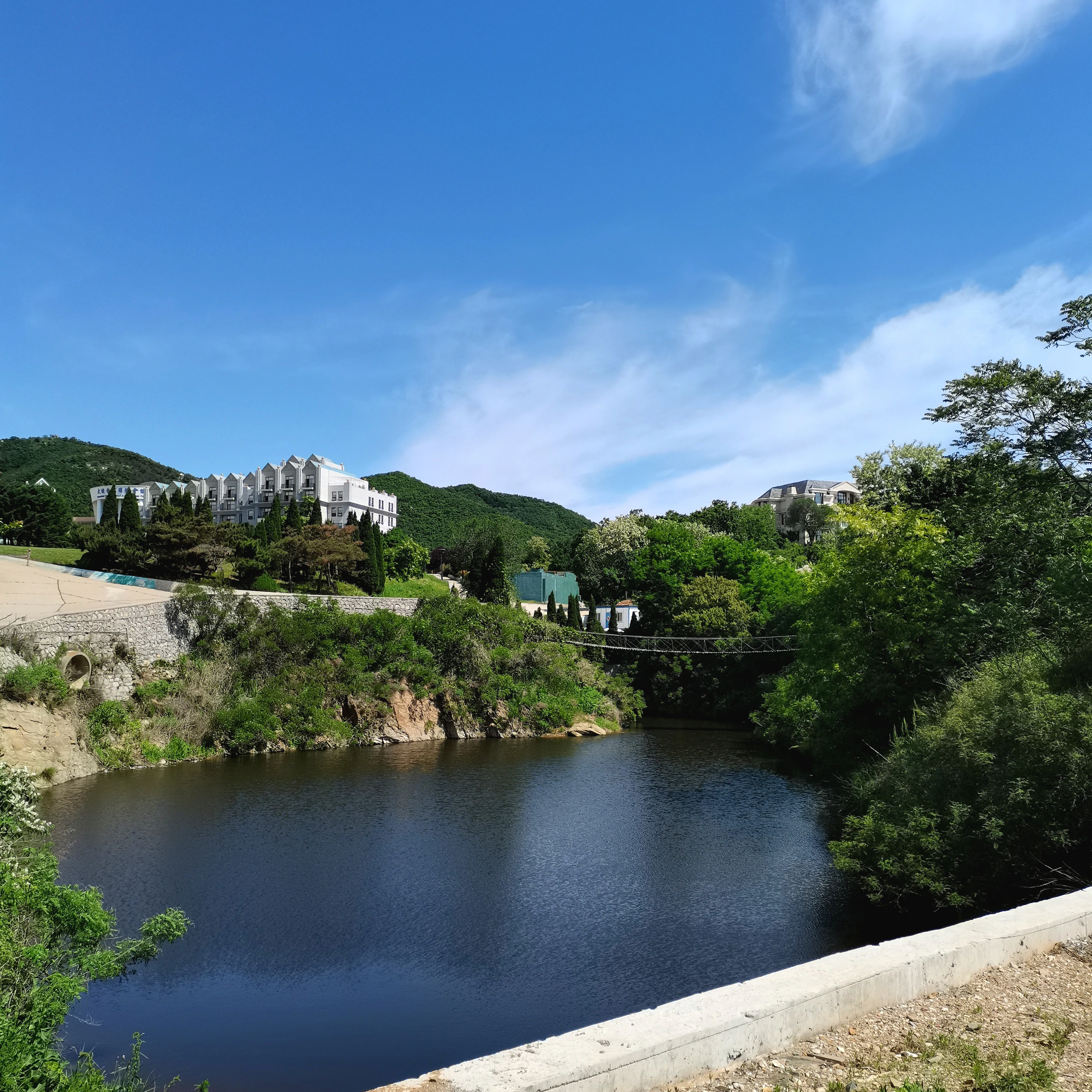 A view across a calm, dark pond toward a forested hill with a white resort building on top and a suspension bridge crossing the water.