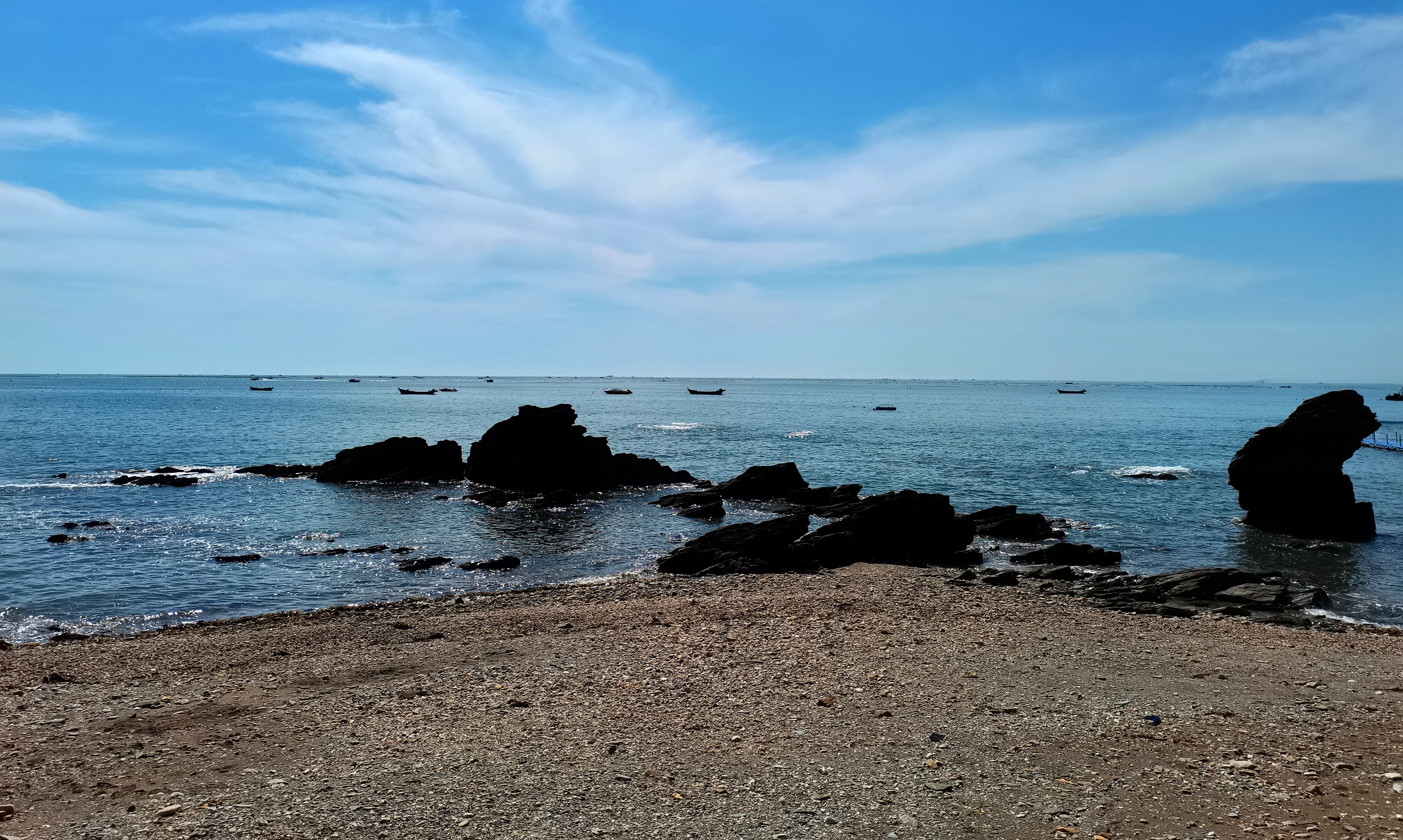 A rocky beach shoreline leading to the ocean under a clear blue sky with thin clouds; small boats are visible in the distant water.