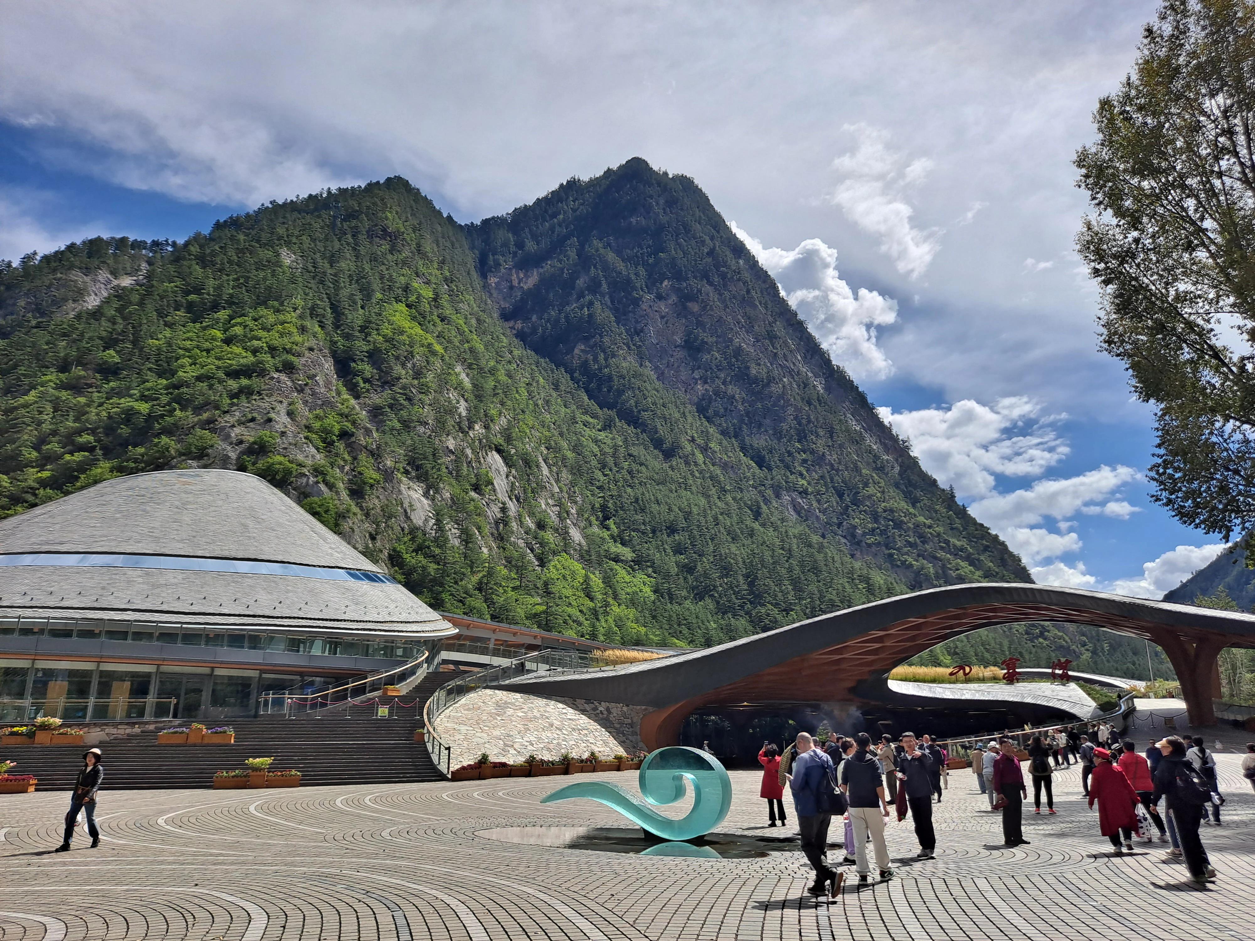 Visitors walking through the paved plaza of the Jiuzhaigou National Park entrance, with a large, modern arched structure and a domed visitor building set against steep, forested mountains.