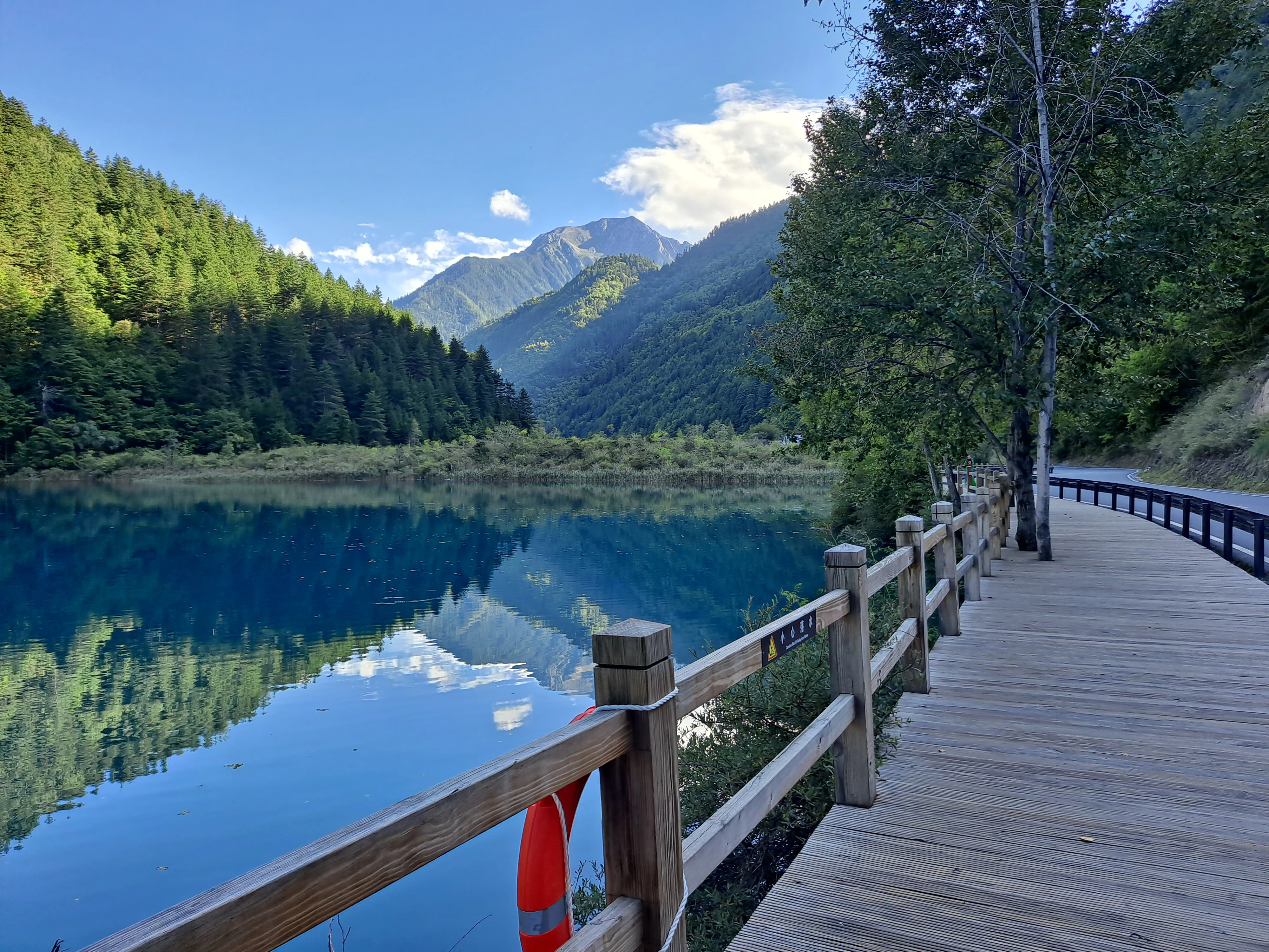 A wooden boardwalk pathway leads along the shore of a vibrant blue lake with mountain slopes and forest in the background.