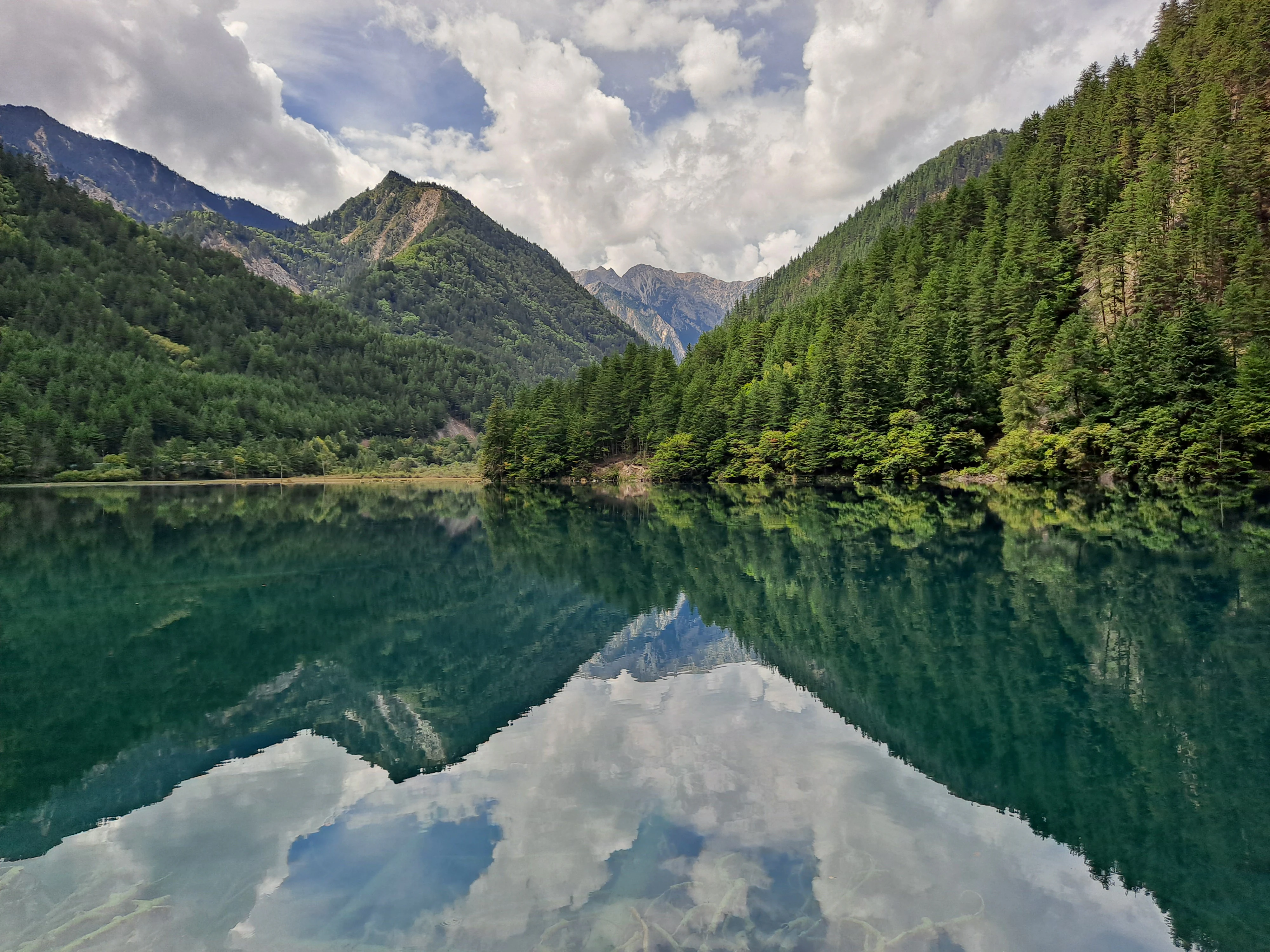 A wide, scenic view of Mirror Lake, where the deep green water creates a near-perfect mirror reflection of the steep, forested mountains and cloudy sky above.