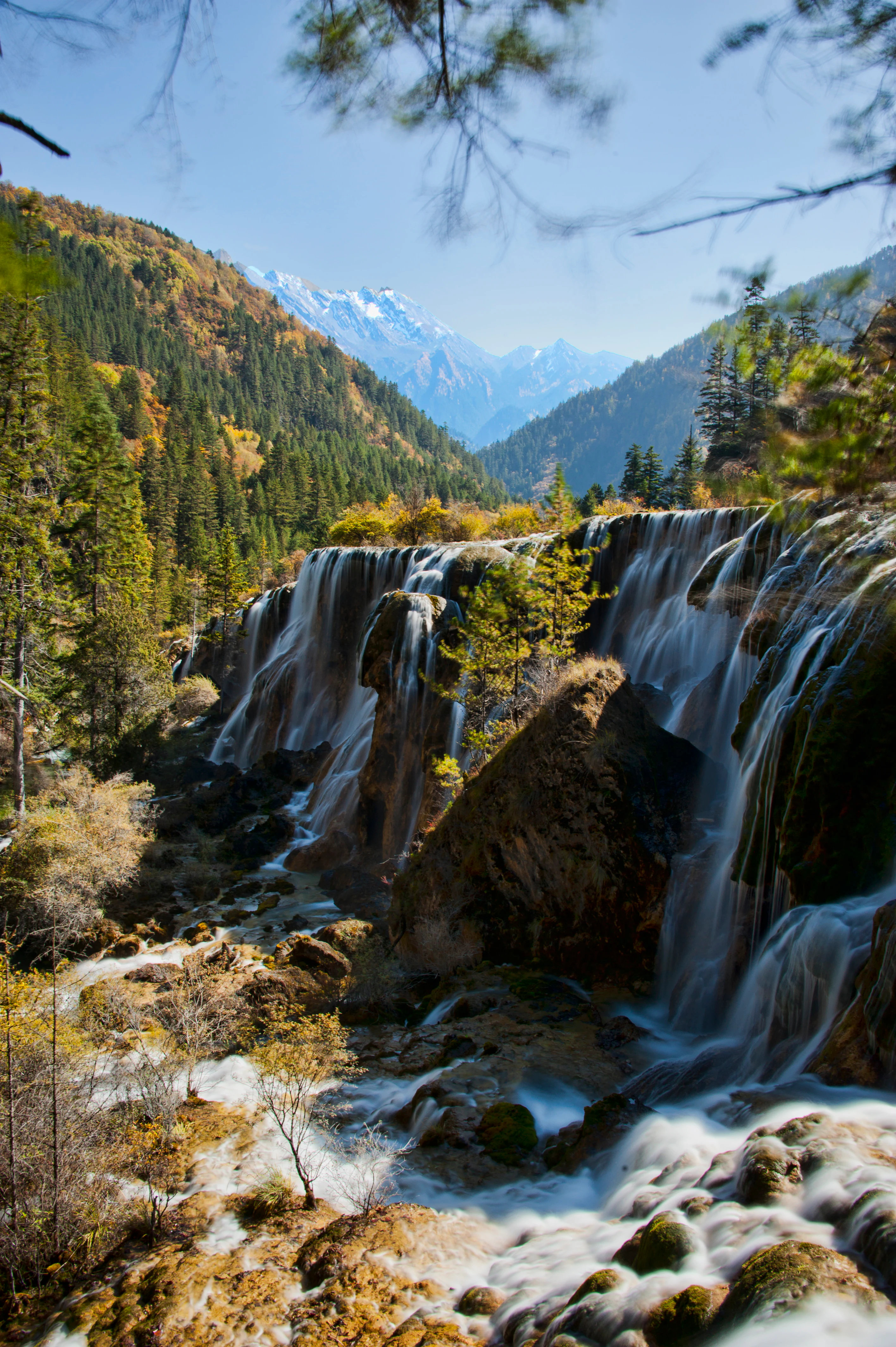 A wide, multi-tiered waterfall flows over rocky, moss-covered ledges, surrounded by a dense coniferous forest in an autumn valley with snow-capped peaks in the background.