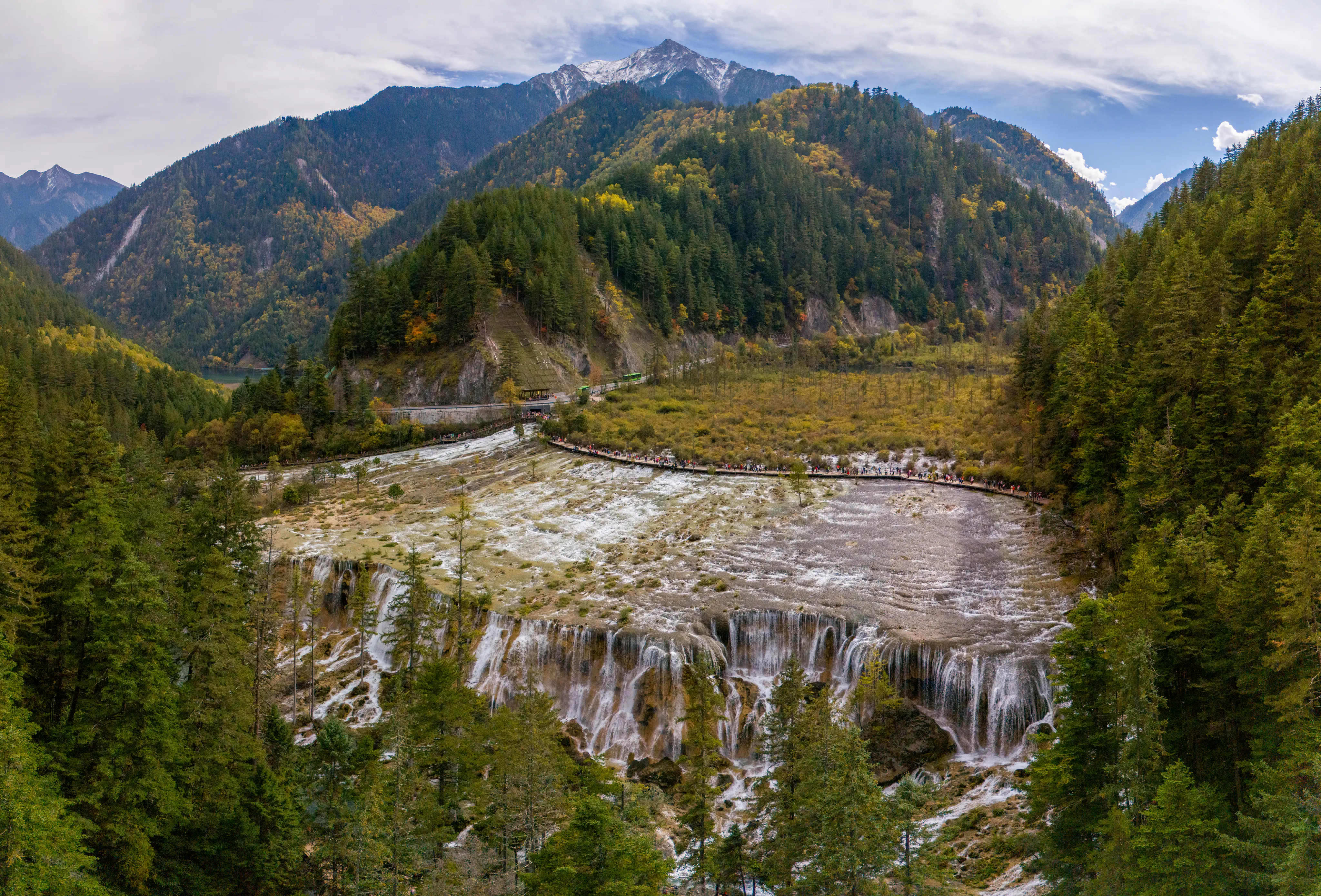 A wide, high-angle landscape shot of Pearl Shoal Falls in Jiuzhaigou, featuring a broad, shallow waterfall cascading over terraced rock steps surrounded by dense coniferous forest and towering, snow-dusted mountains under a cloudy sky.