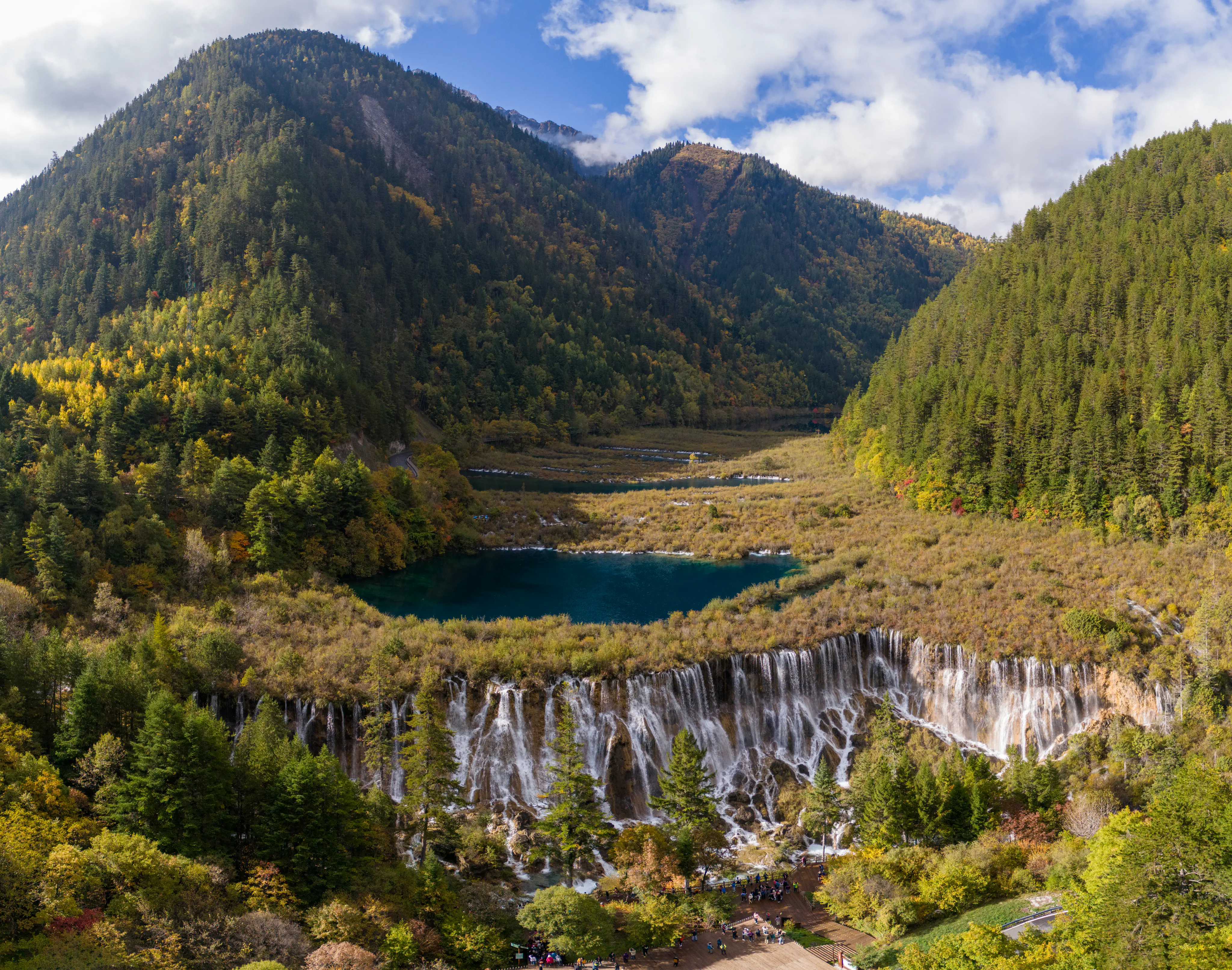 An elevated, wide-angle view of the iconic Nuorilang Waterfall flowing from a deep blue-green lake, set within a lush, forested valley in the mountains during autumn.