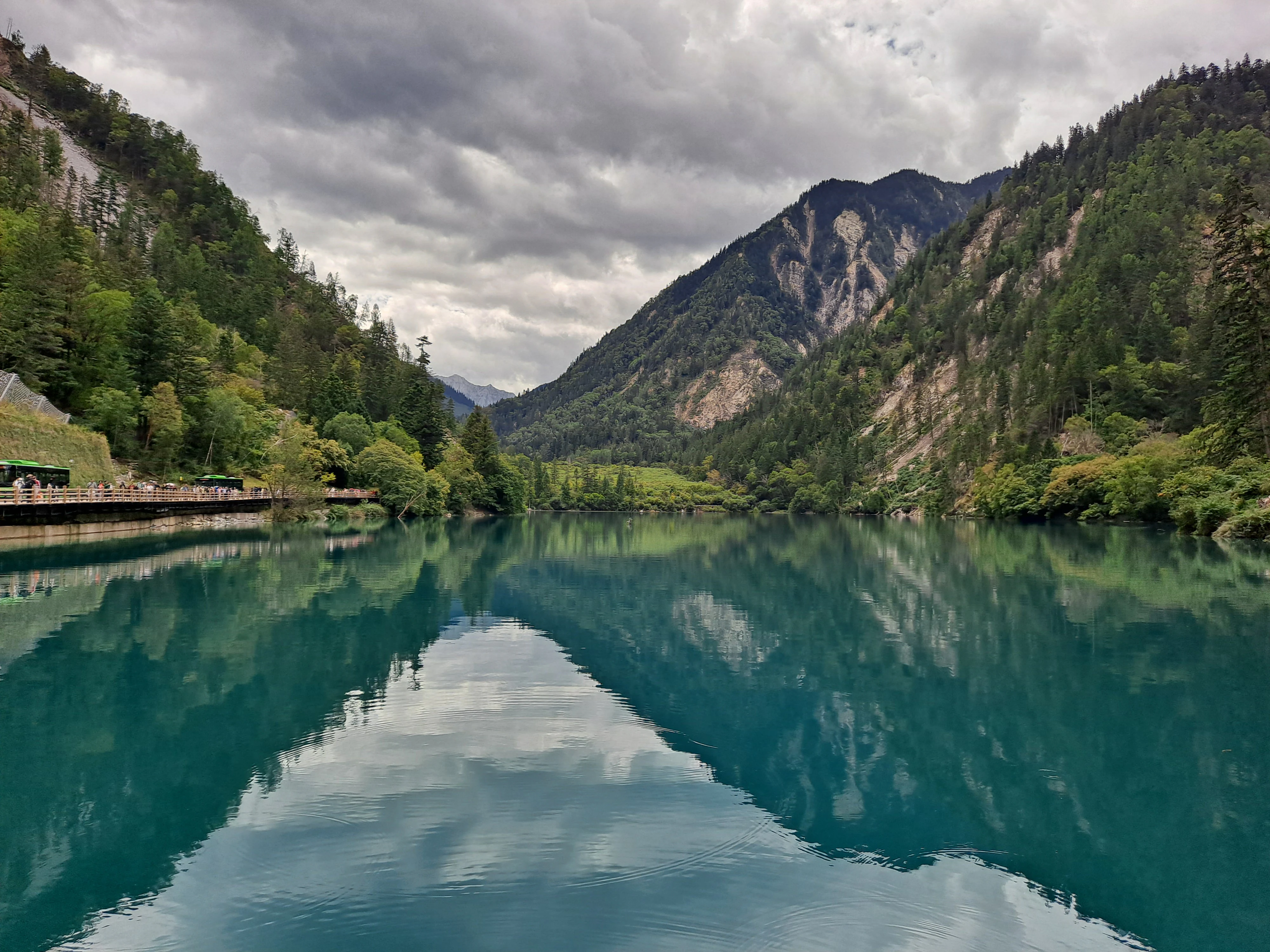 A wide shot of a calm, turquoise lake surrounded by steep, forested mountains under a cloudy sky; a wooden boardwalk is visible on the left bank.