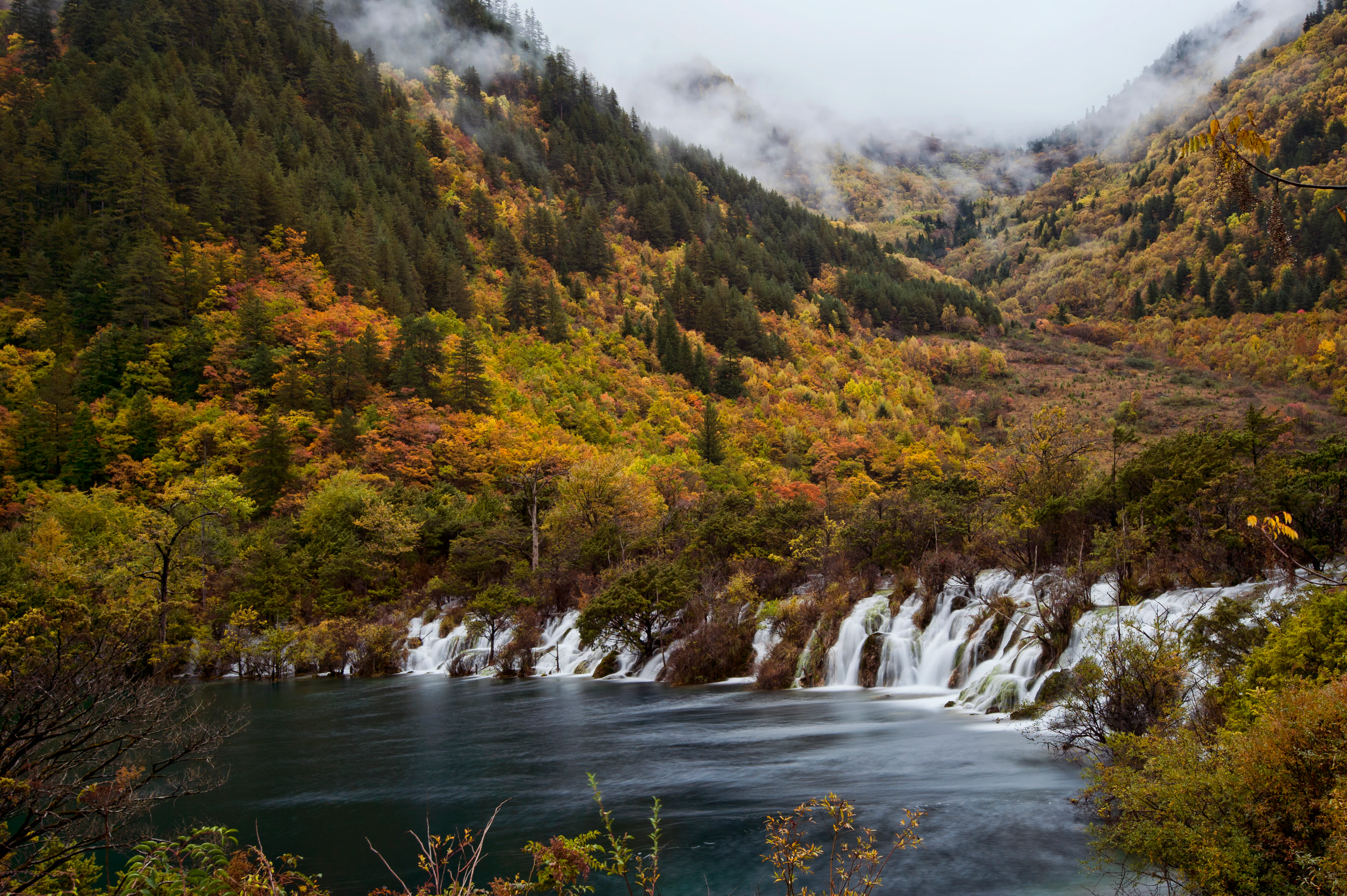 A wide view of a multi-tiered waterfall cascading into a calm turquoise lake, surrounded by forested mountains displaying autumn foliage and misty peaks.