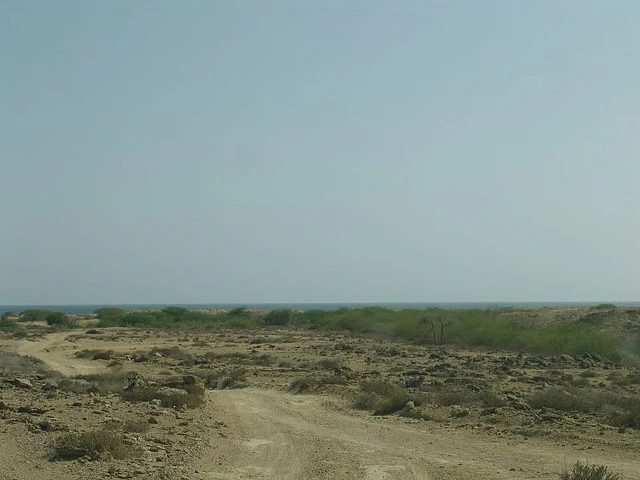 Una toma amplia de un camino polvoriento y sin pavimentar que serpentea por un paisaje costero árido y rocoso bajo un cielo brumoso, con el océano azul oscuro visible en el horizonte lejano.