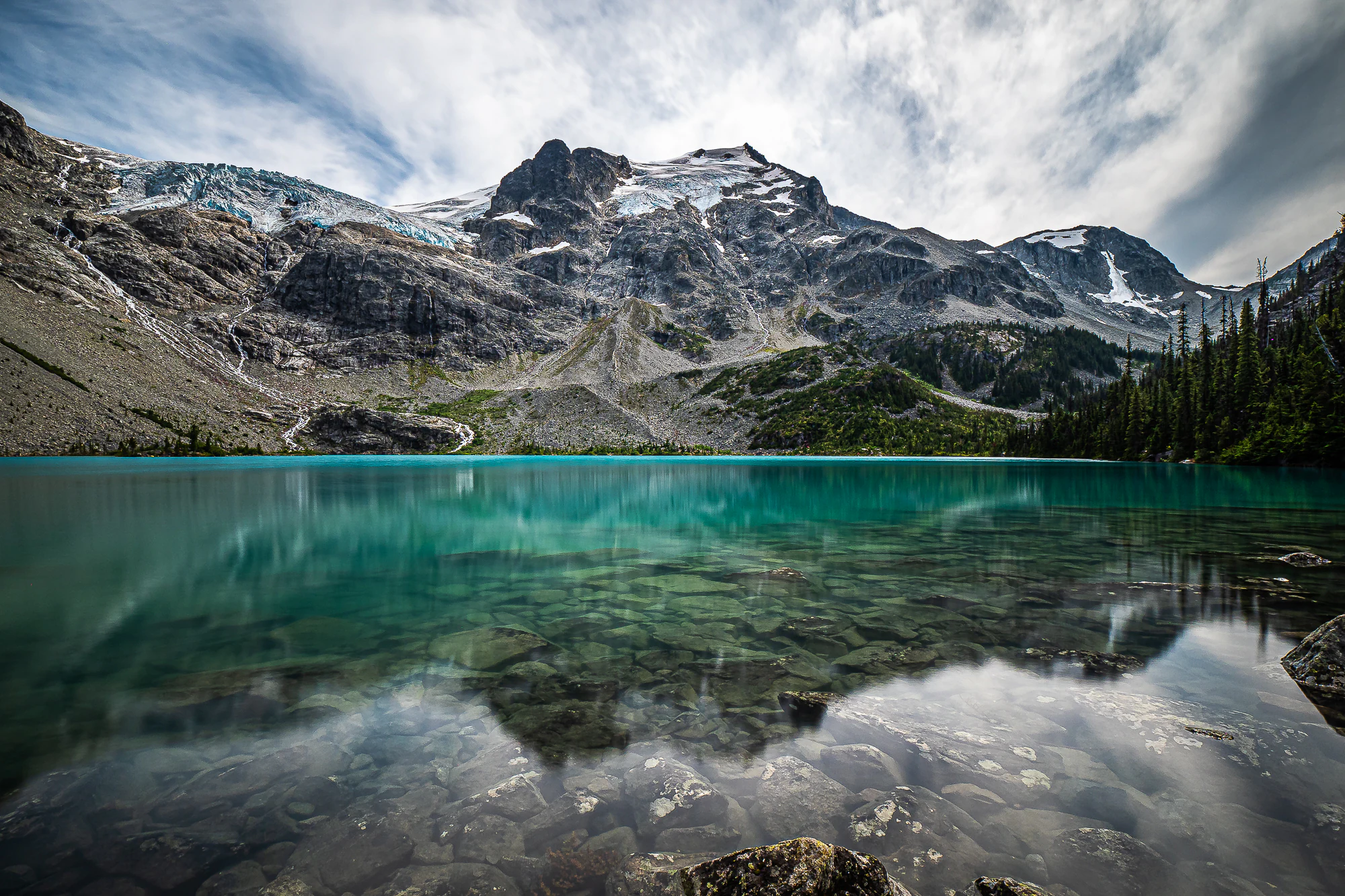 A wide angle view of a bright turquoise lake in the foreground, with clear water revealing submerged rocks, and a massive, snow-dusted, craggy mountain peak with a visible glacier in the background under a cloudy sky.