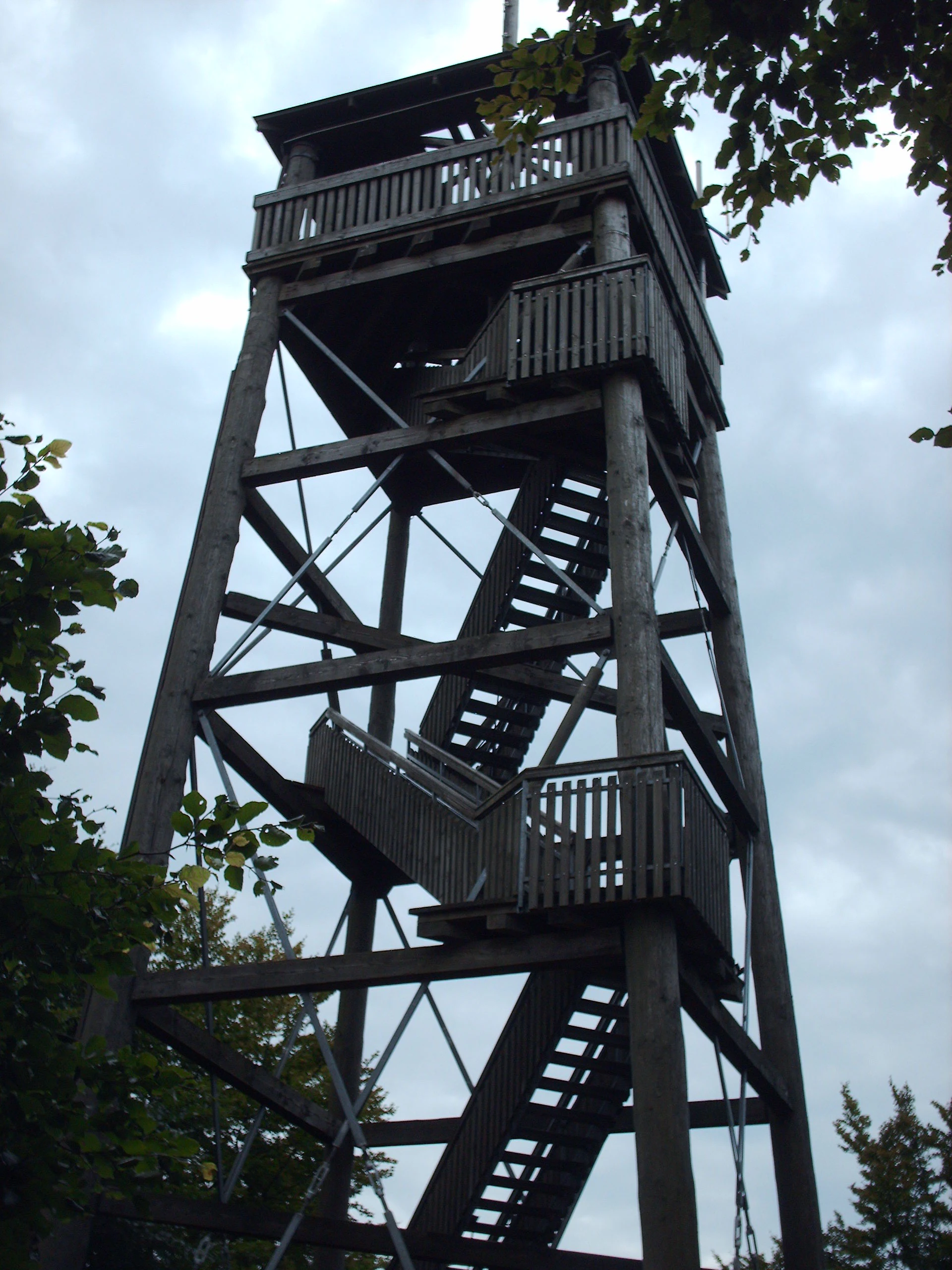 A tall, wooden lattice-style observation tower with an external staircase rising against a cloudy sky.