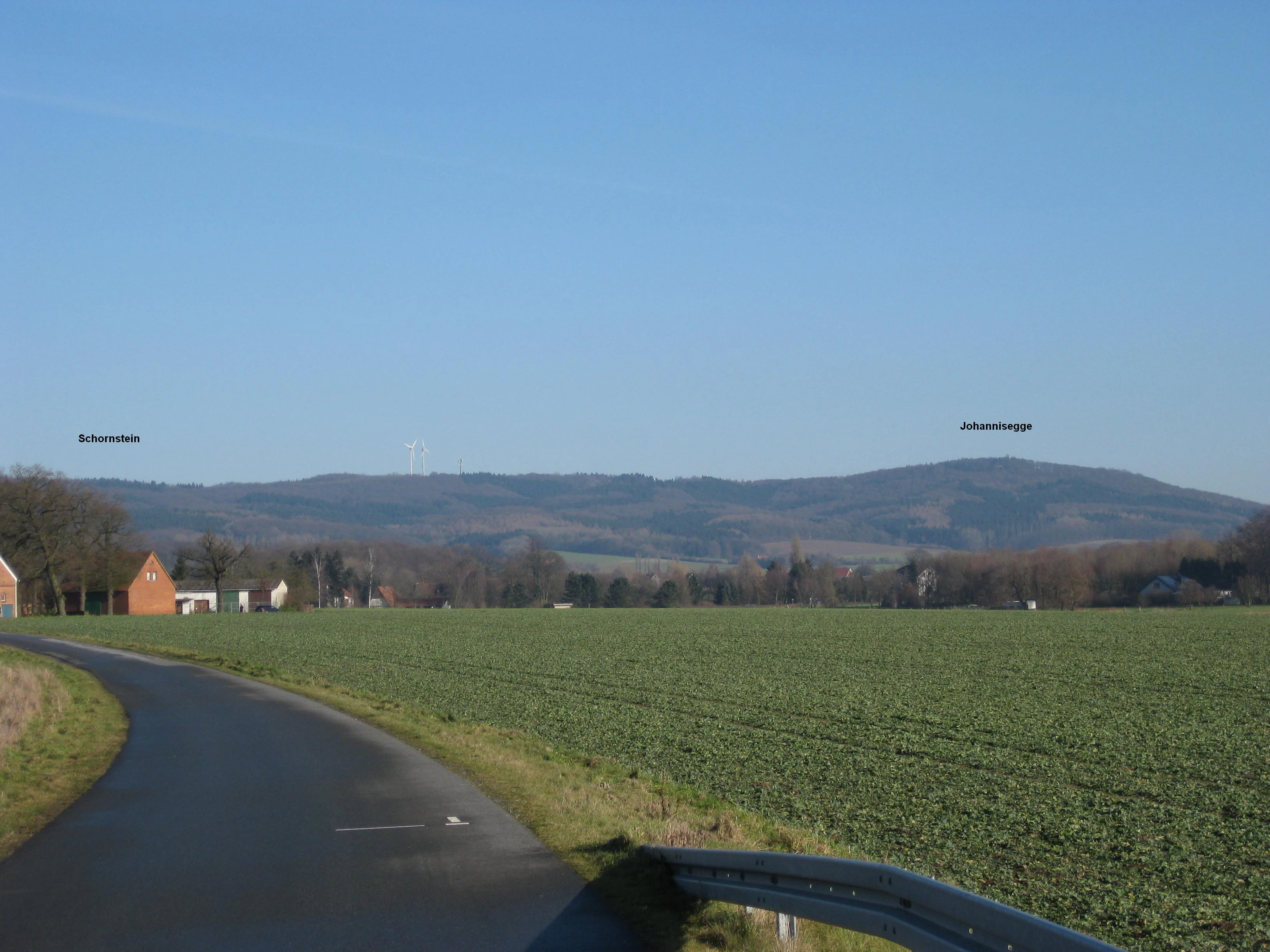 A wide, eye-level outdoor shot showing a green agricultural field and an asphalt road in the foreground, with distant, low rolling hills covered in forest under a clear blue sky. Small text labels identifying Johannisegge and Schornstein are visible on the horizon.