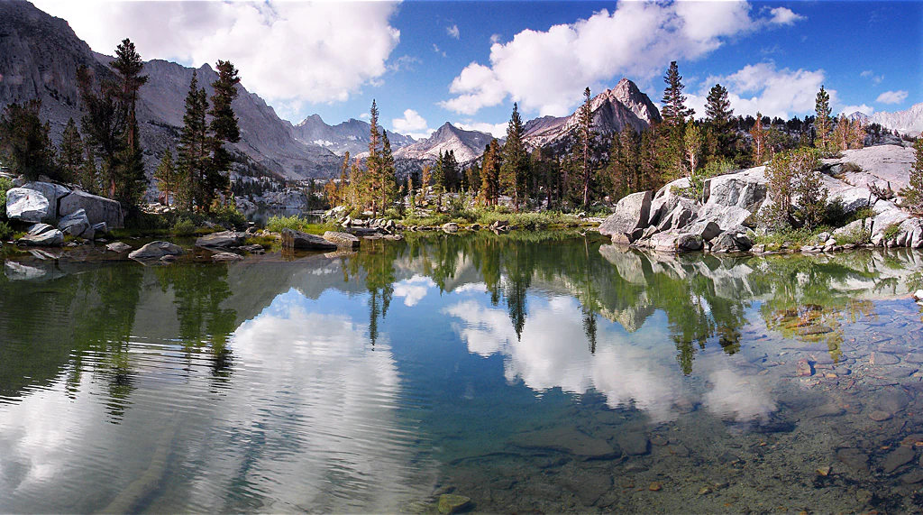 A calm, clear lake with rocky shores and pine trees reflects the surrounding jagged Sierra Nevada mountain peaks and a blue sky with white clouds.