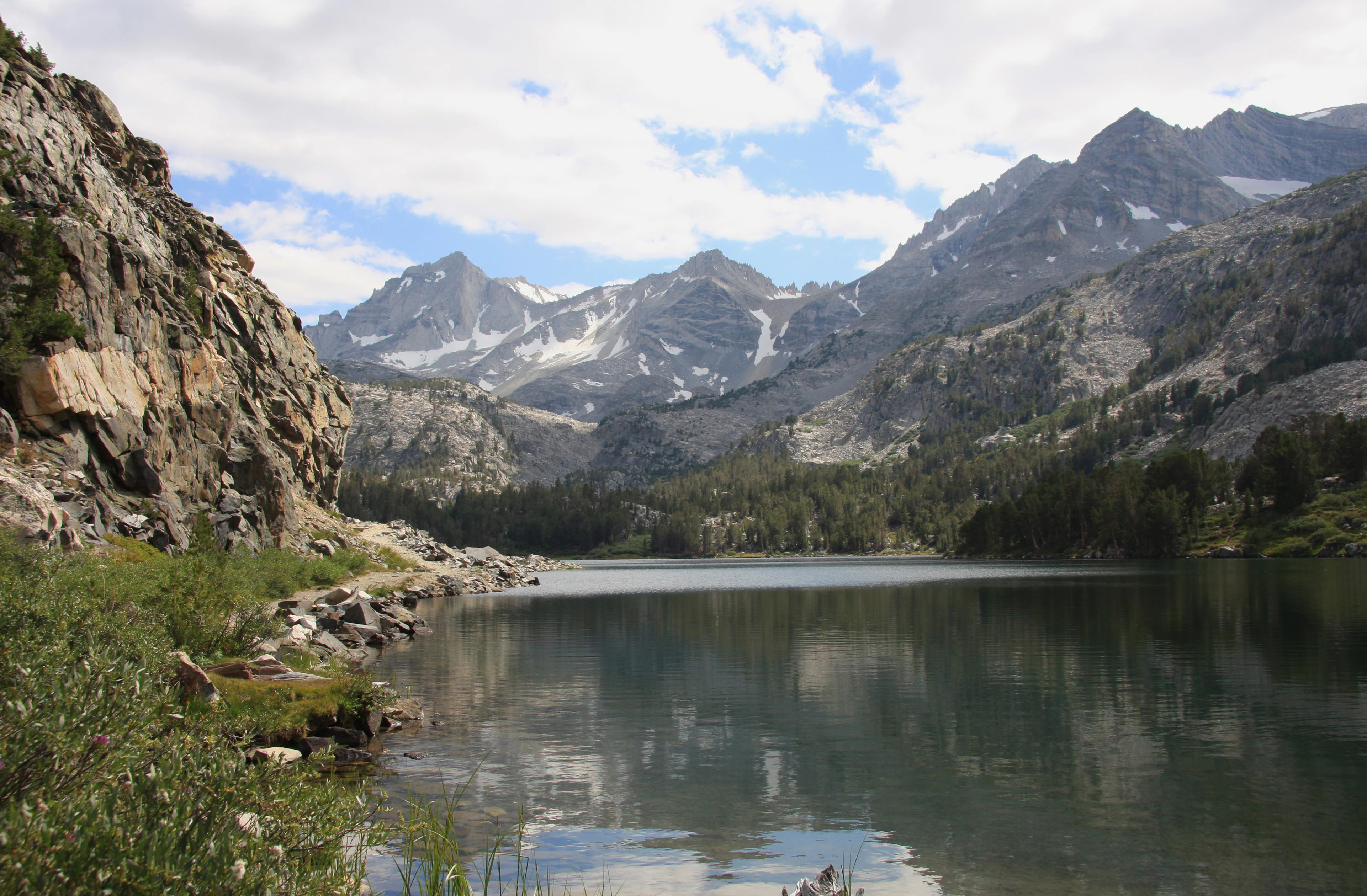 An alpine lake with calm, reflective water surrounded by rocky cliffs, pine trees, and snow-capped peaks in the background under a partly cloudy sky.