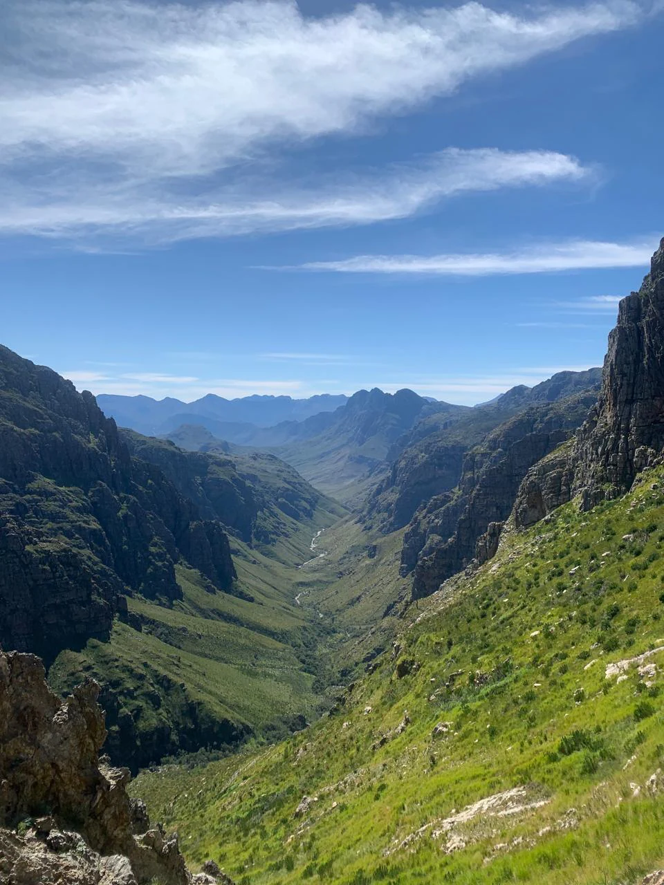 A high angle view looking down into a deep, green mountain valley with steep rocky slopes on both sides under a bright blue sky with wispy clouds.