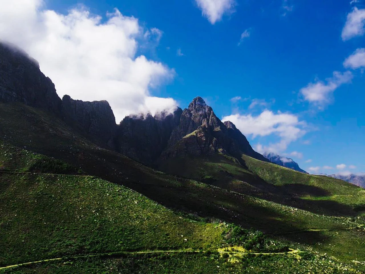 A wide angle view of a jagged mountain range in Jonkershoek Nature Reserve, with green grassy slopes in the foreground and a blue sky with scattered clouds above.