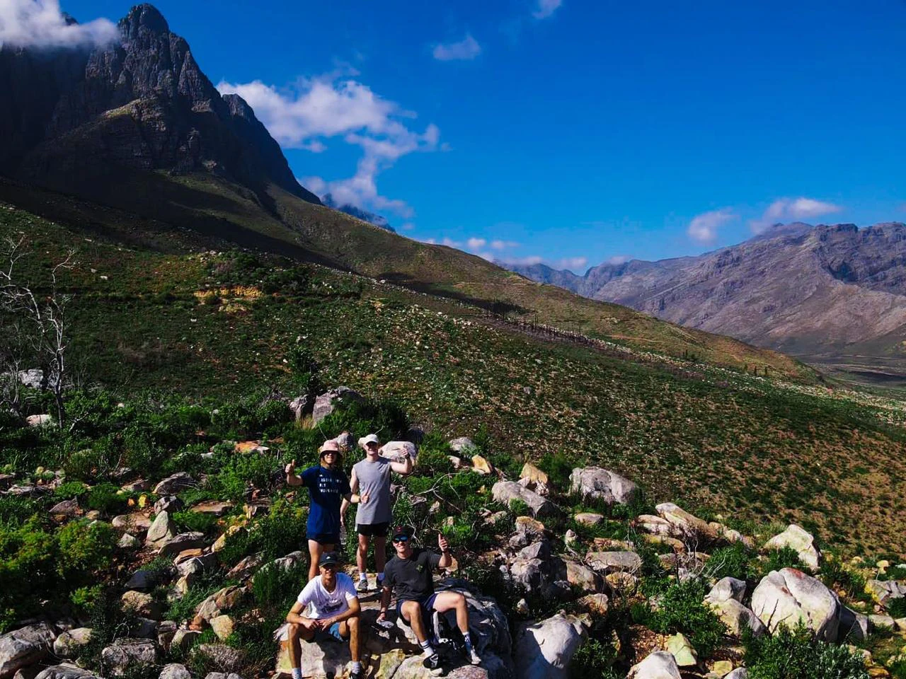 A group of five people sitting and standing on rocks along a hiking trail, with large mountain peaks under a clear blue sky in the background.