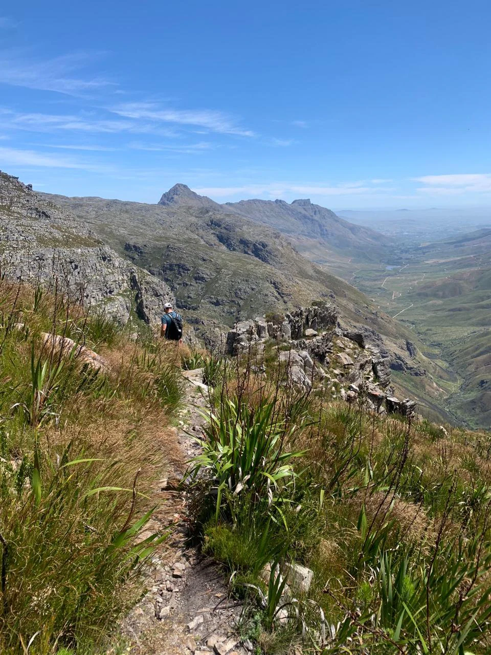 A hiker with a backpack walks along a rocky, narrow trail on a mountainside, with a vast mountain valley visible in the background under a blue sky.