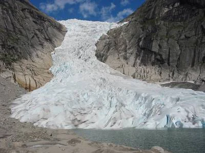 A large glacier descending between rocky mountain slopes with a body of water at its terminus under a partly cloudy sky