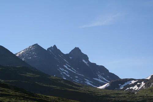 A mountain landscape with rocky peaks, patches of snow, and green slopes under a clear blue sky