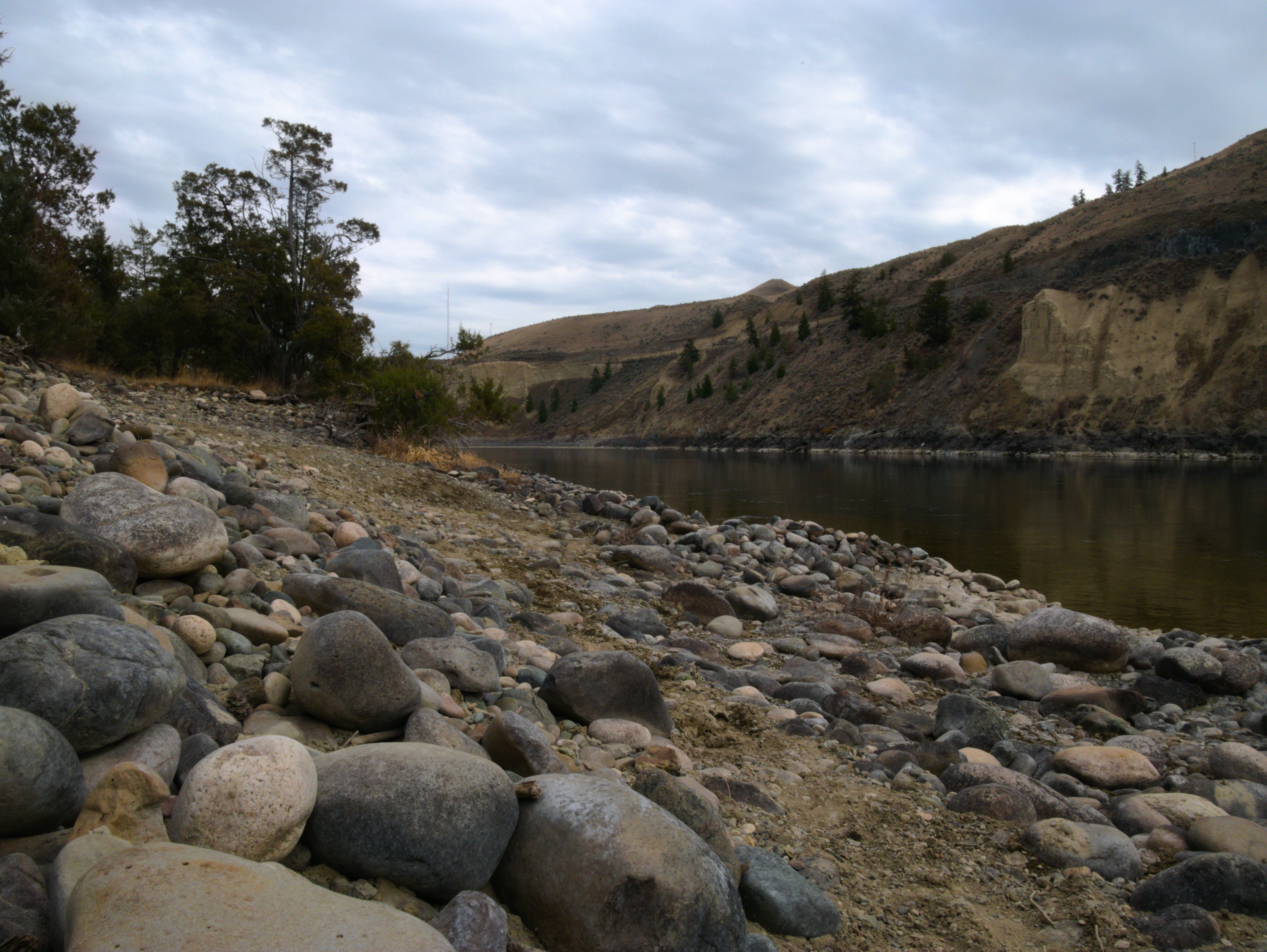 A rocky, dry riverbank with smooth stones in the foreground; the Thompson River flows calmly in the midground, flanked by rolling, arid hills under a cloudy sky.